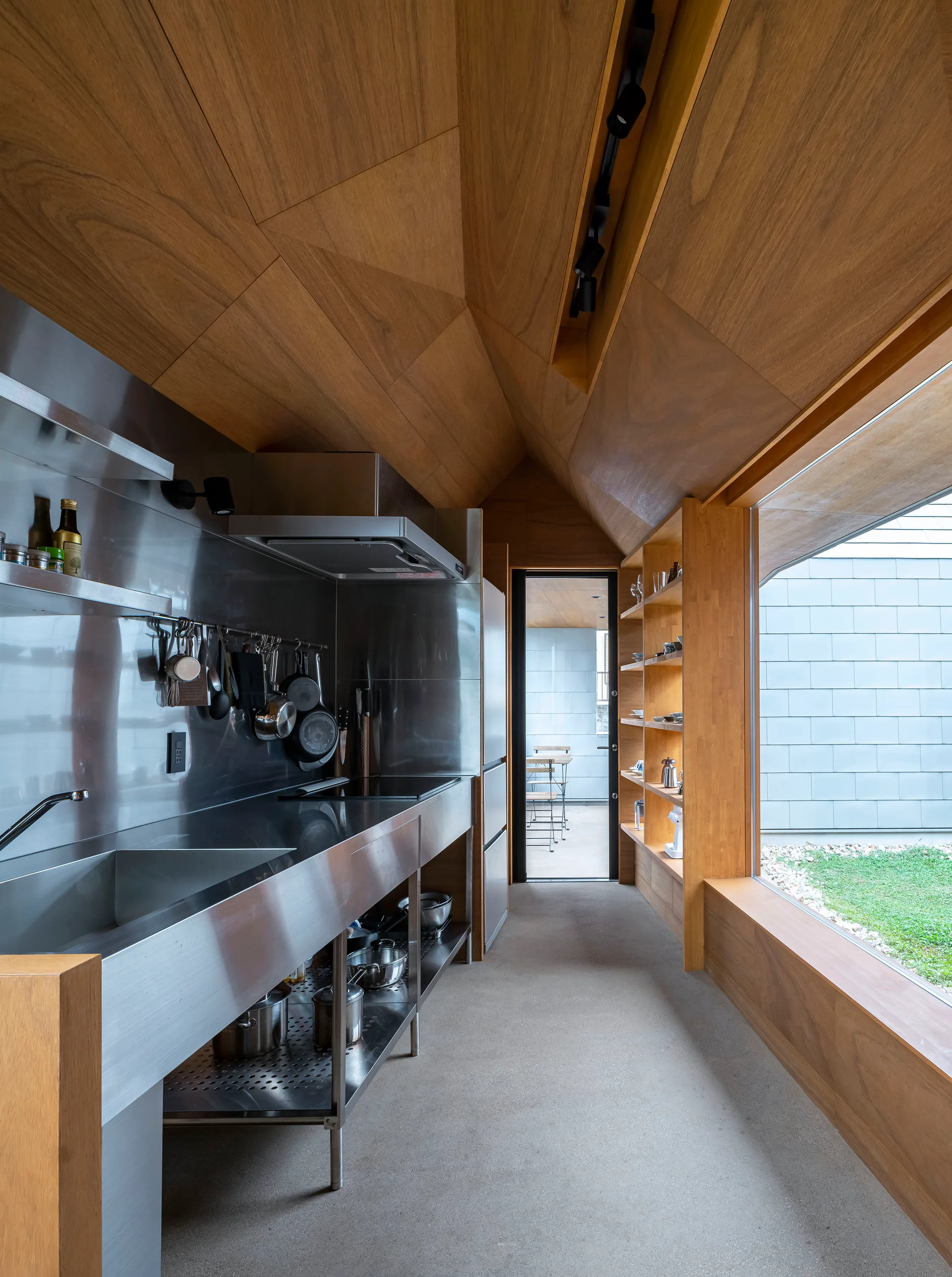 A kitchen with a long stainless steel counter, vaulted wood paneled ceiling, concrete floor, and a picture window overlooking a yard.