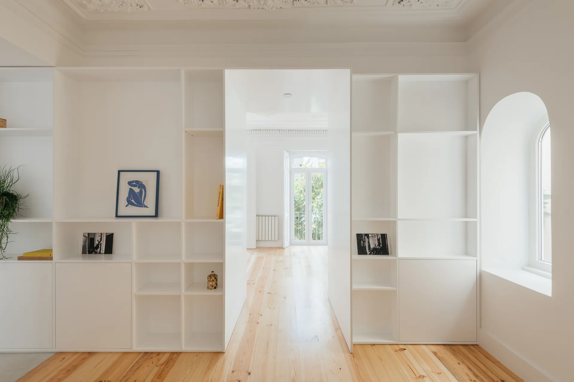 White floor-to-ceiling built-in bookshelves frame an open central passageway in a room with light wood floors and an arched window.
