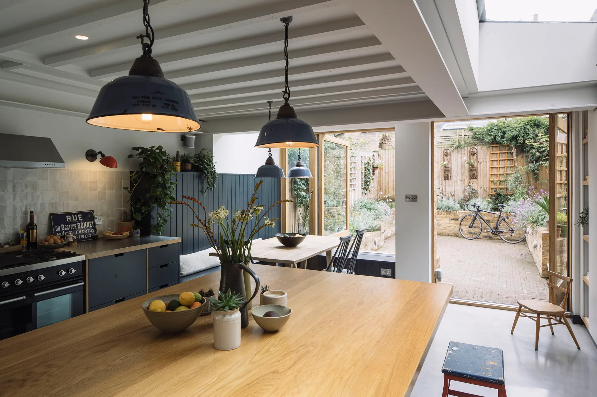 A modern kitchen featuring a large oak island, navy blue cabinets, white ceiling beams, and bifold doors opening to an outdoor courtyard.