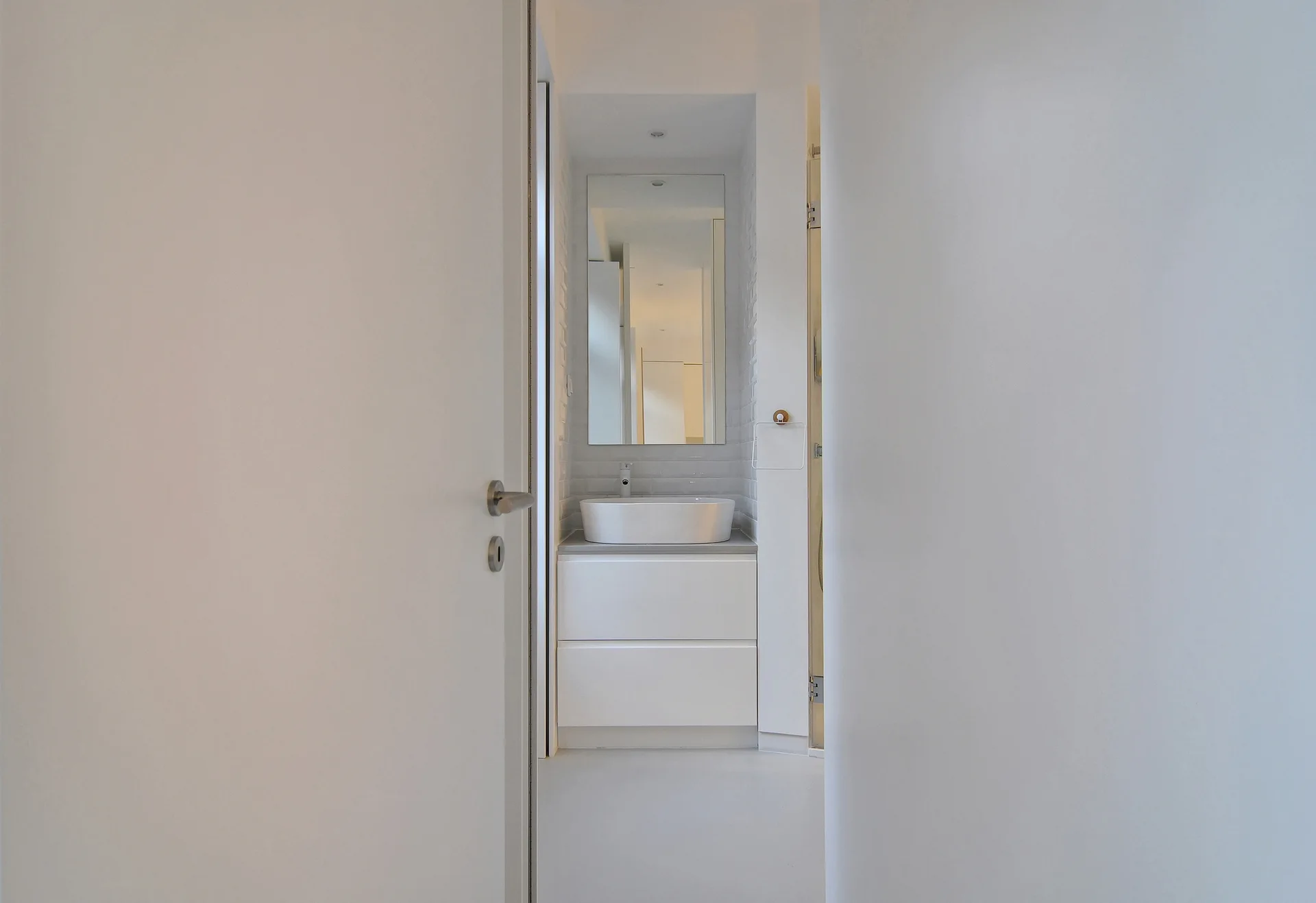 A view through a white doorway into a minimalist bathroom featuring a white vanity cabinet with a vessel sink and a rectangular mirror.