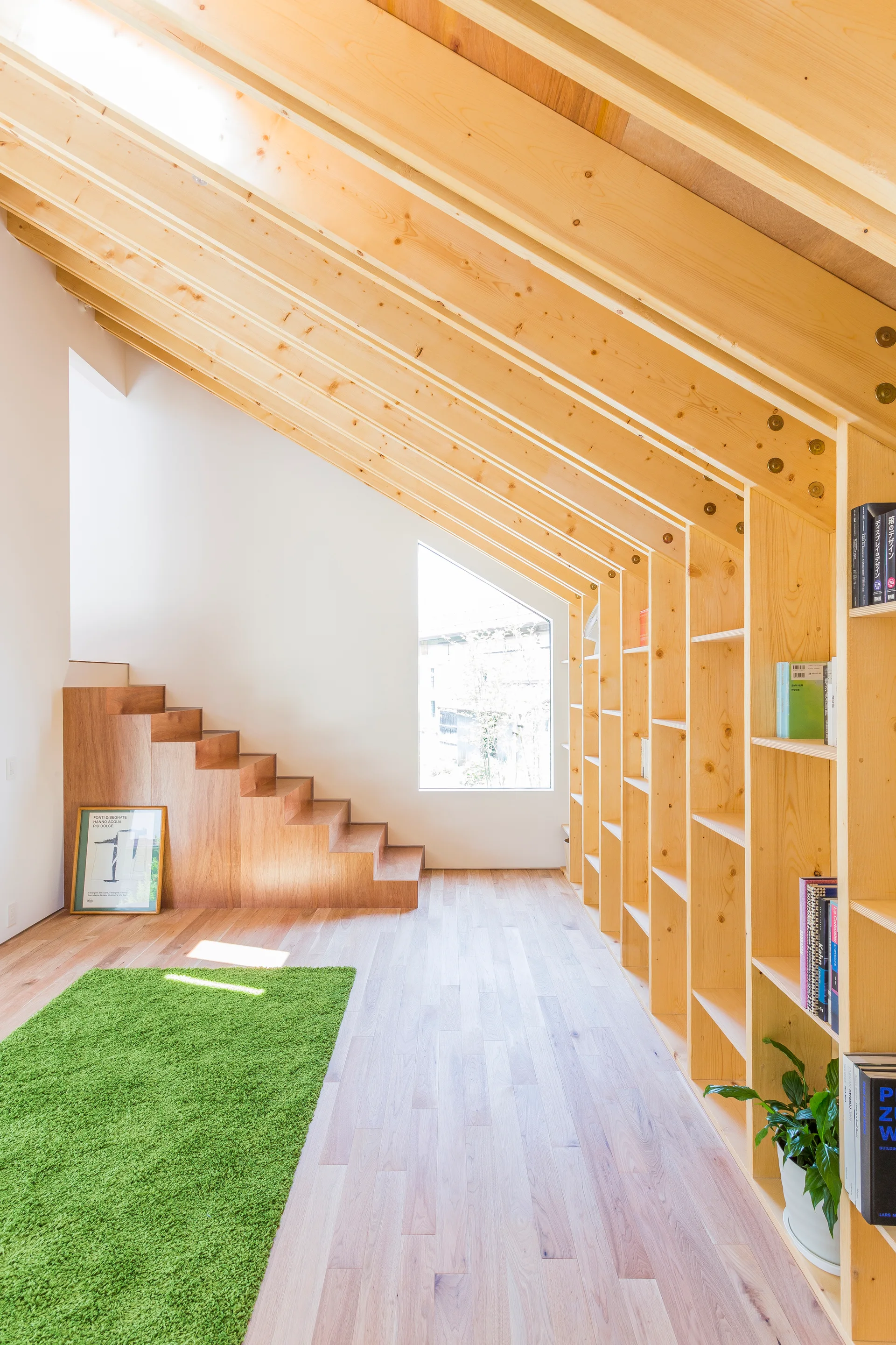 A bright loft space featuring sloped wooden rafters, integrated pine bookshelves, a built-in staircase, and a vibrant green rug on hardwood flooring.