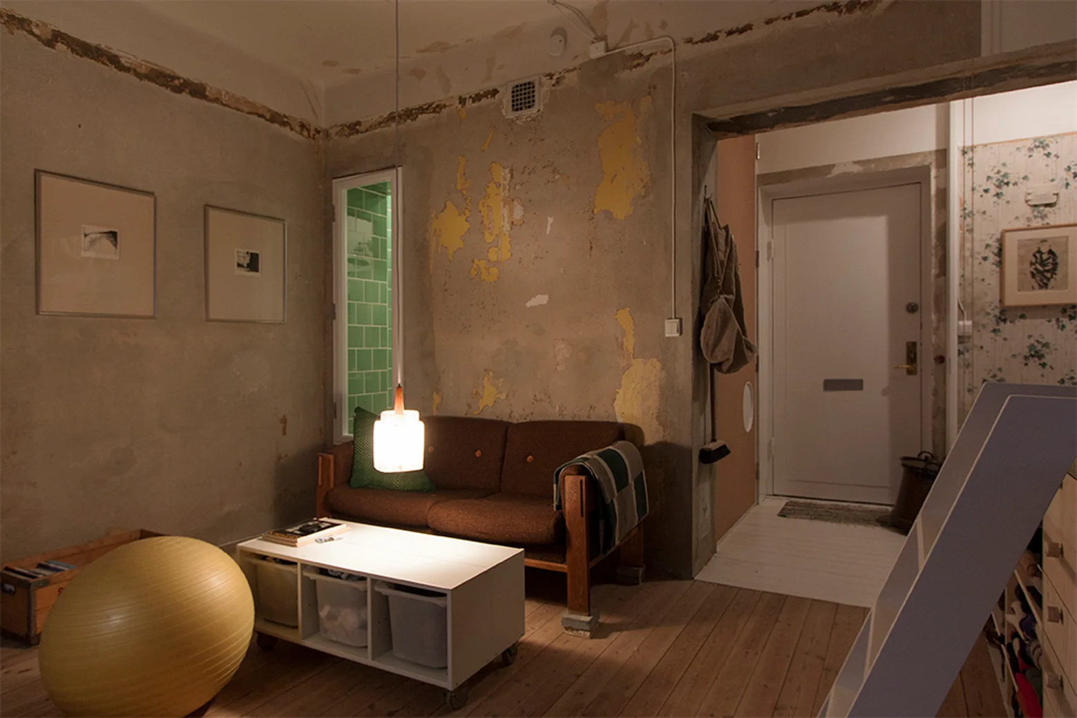 A casual living room features textured, unfinished plaster walls, a brown sofa, a low white coffee table, and a yellow exercise ball on wood floors.