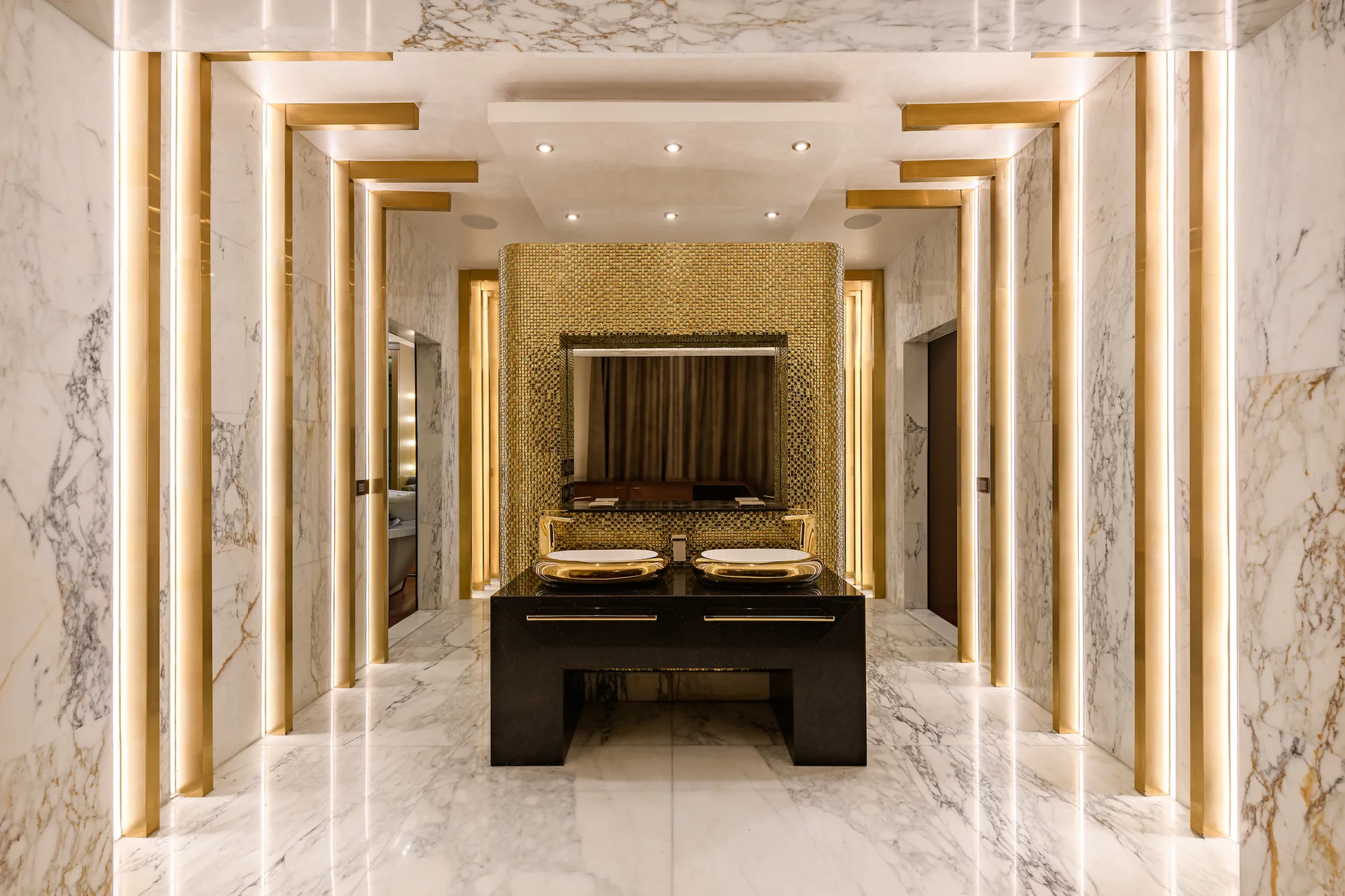 Symmetrical bathroom featuring a black double vanity with gold sinks against a gold mosaic wall, flanked by marble columns with glowing light strips.