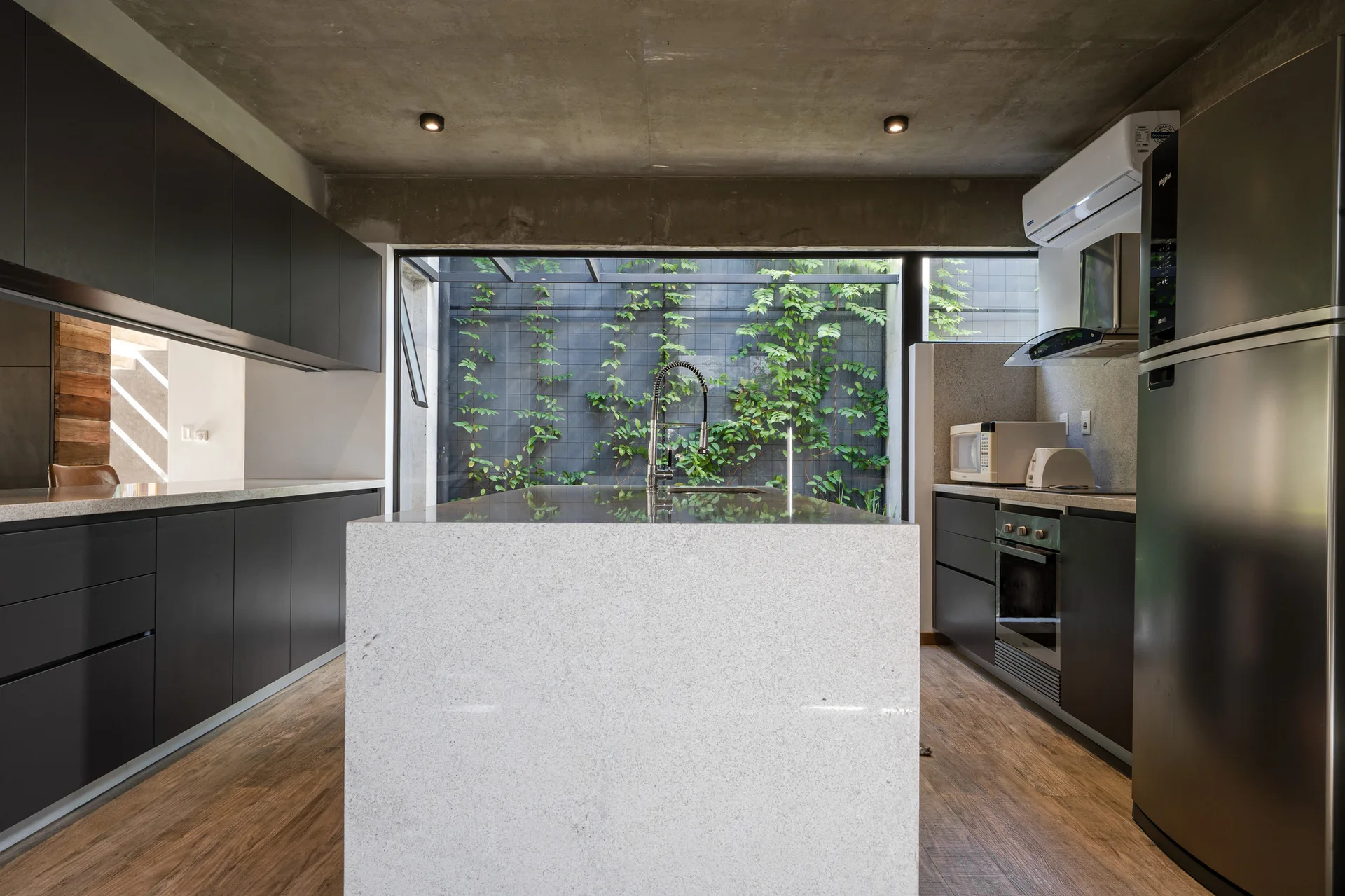 A modern kitchen featuring a light stone island, matte black cabinets, an exposed concrete ceiling, and a large window overlooking climbing green ivy.