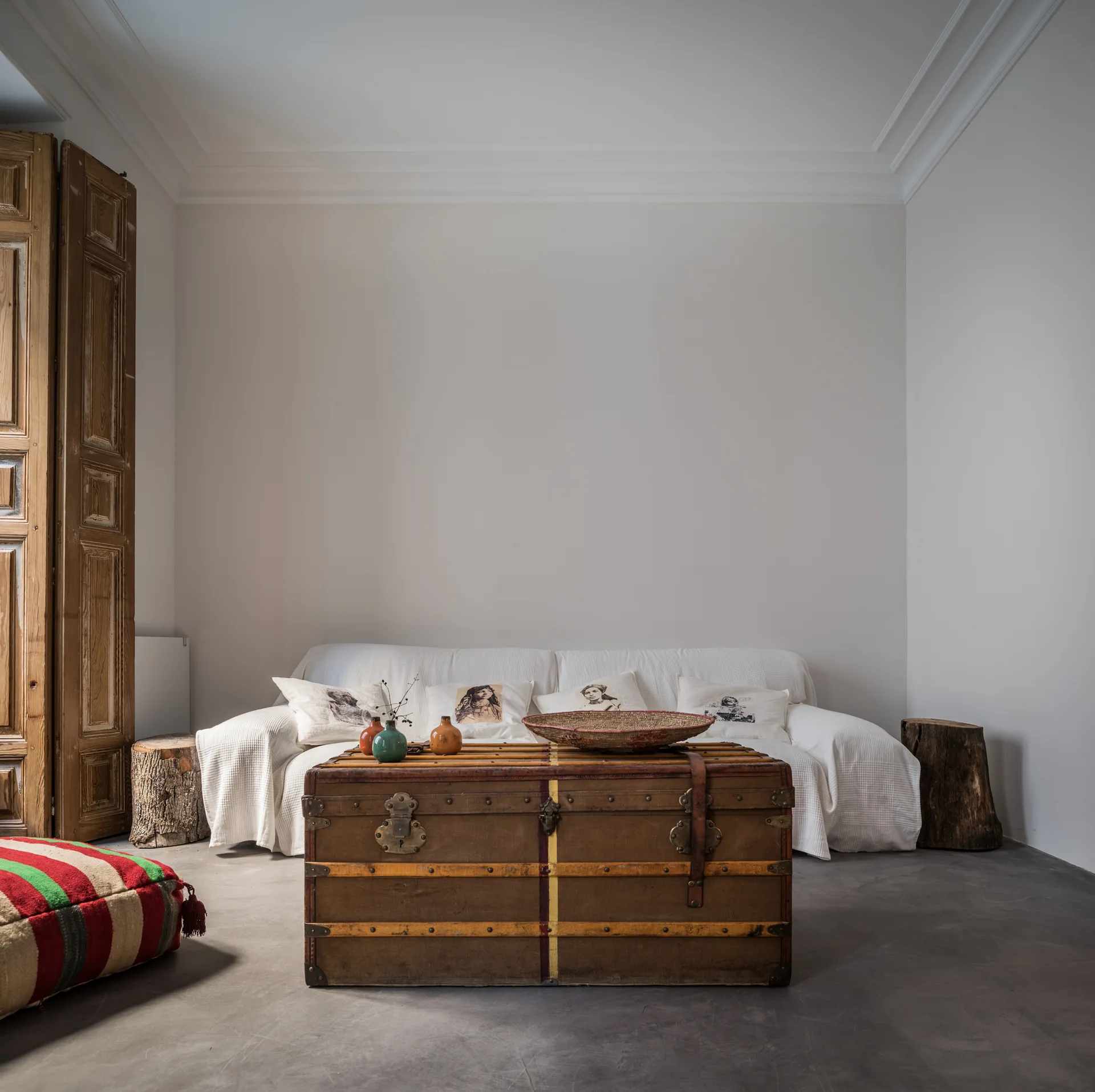 A white sofa rests against a plain wall behind a vintage wooden trunk coffee table on concrete flooring with log stools.