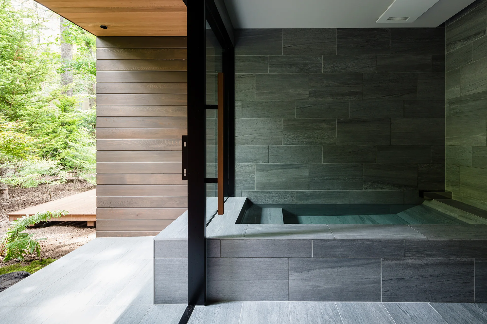 A modern grey stone soaking tub is built into a tiled room next to a sliding glass door opening to a wooden deck and green trees.