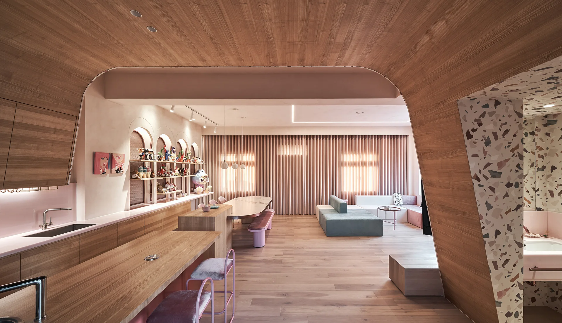 A modern open-concept living area features a curved wood-clad ceiling archway, light pink walls, a long dining table, and modular seating on wood floors.