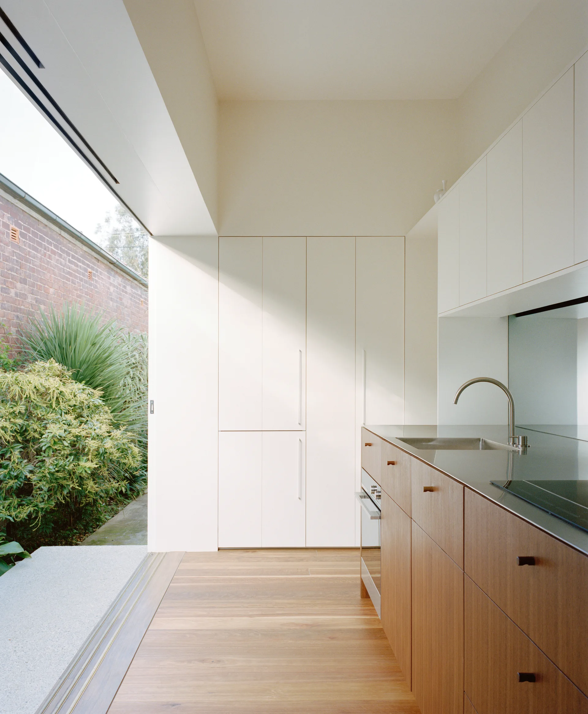 A modern kitchen featuring warm wood base cabinets, matte white tall storage, and a large sliding glass door opening to a lush garden.