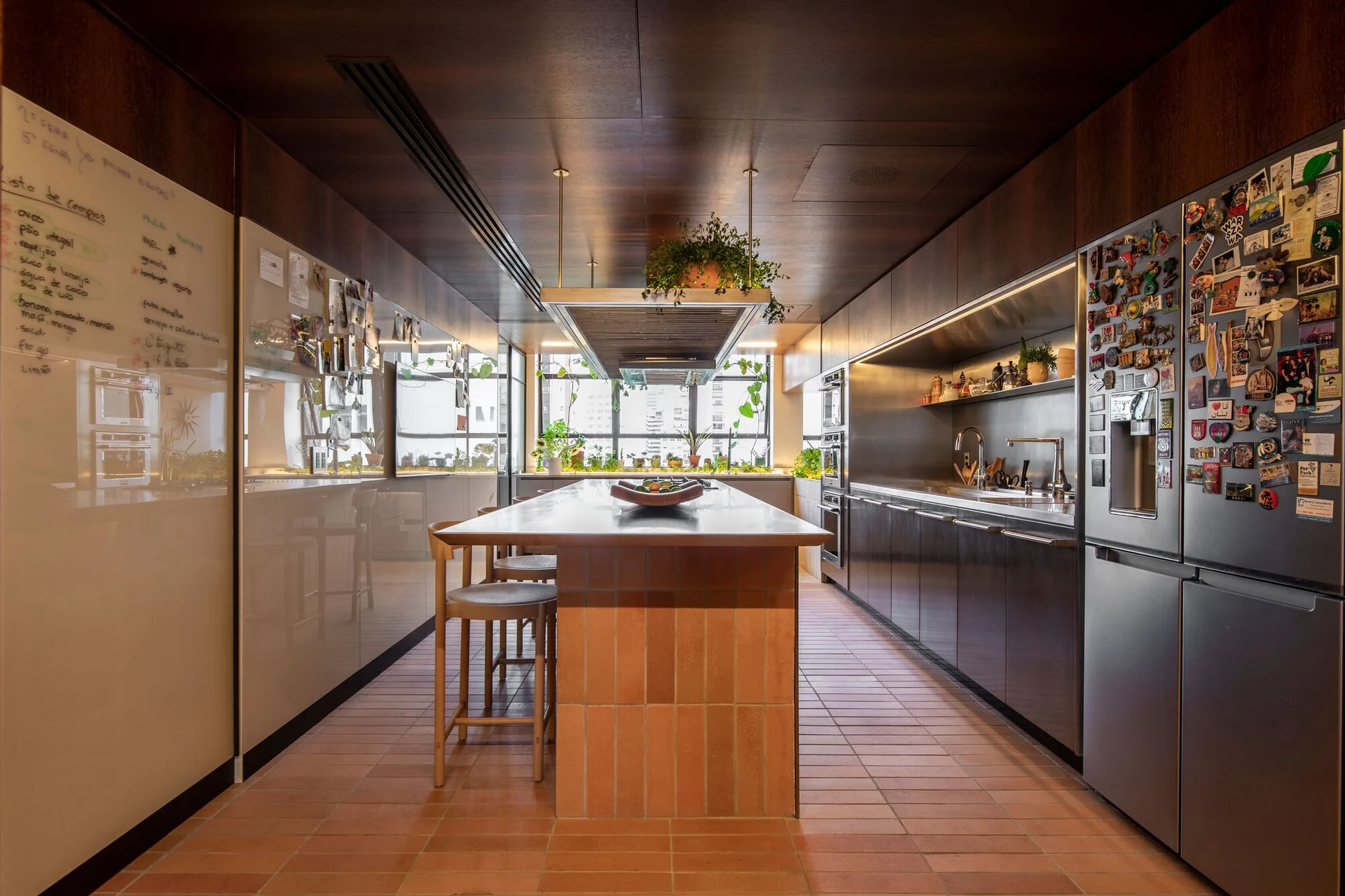 A galley kitchen features a central island clad in terracotta tile, matching floor tiles, dark wood ceiling paneling, and stainless steel appliances.