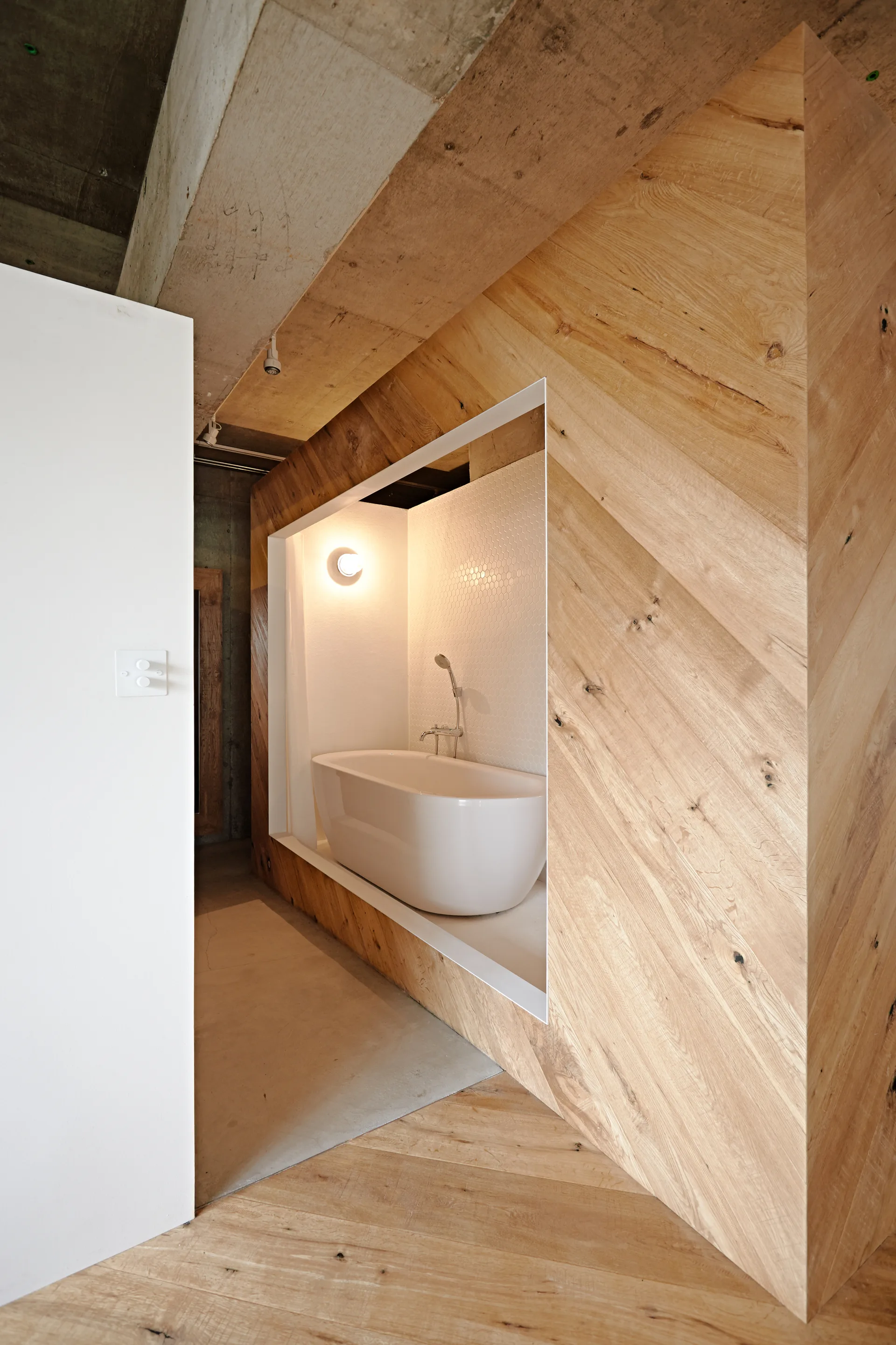 A modern white freestanding bathtub sits inside a tiled niche recessed within a diagonal wood-paneled wall next to an exposed concrete ceiling.