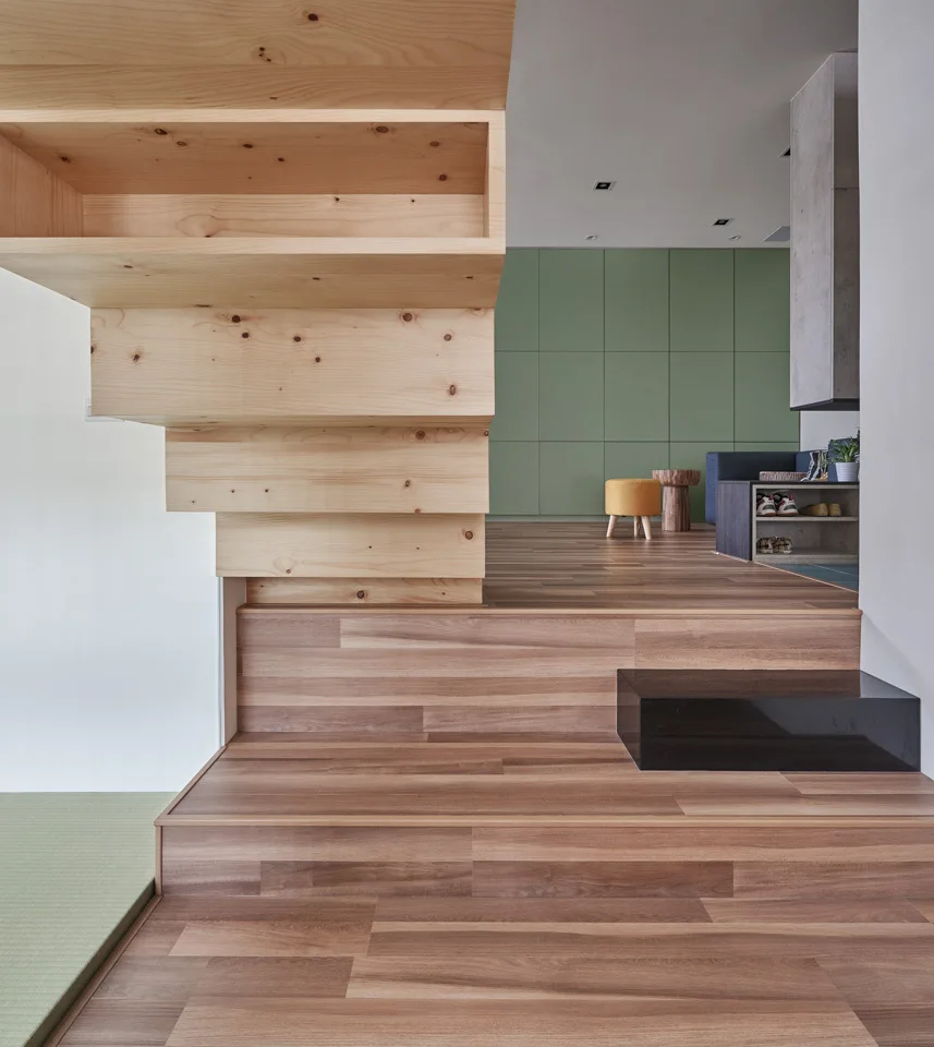 A light wood staircase ascends from a split-level landing with wooden floors toward a background living area featuring a sage green paneled wall.