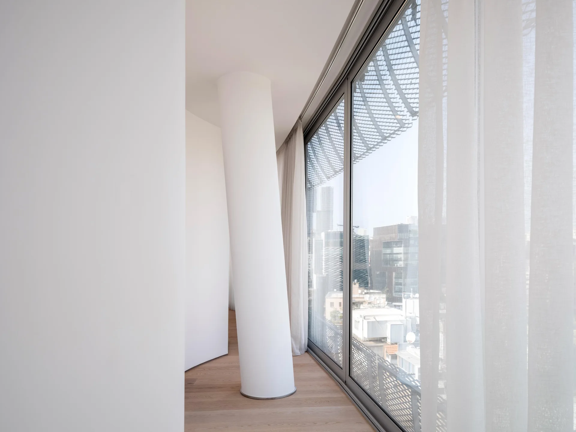 An angled white structural column stands near a large floor-to-ceiling window overlooking a city skyline in a minimalist room with light wood floors.