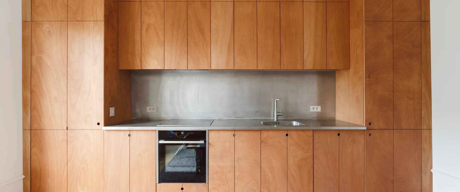 A minimalist kitchen features seamless wood-veneered cabinetry, an integrated oven, and a stainless steel countertop and backsplash.