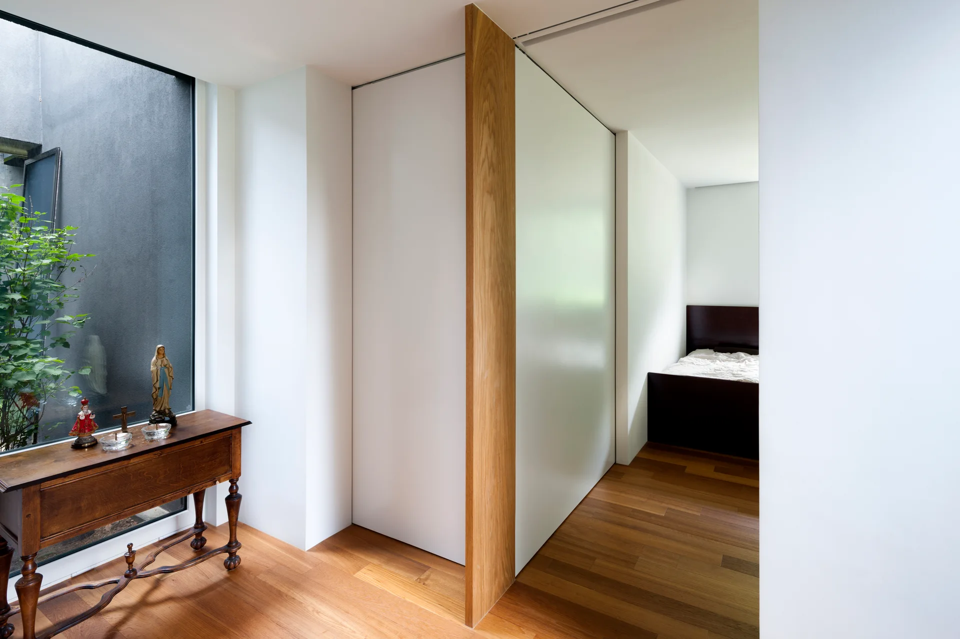 A minimalist hallway with hardwood floors features an ornate wooden console table by a window and a vertical wood partition leading to a bedroom.