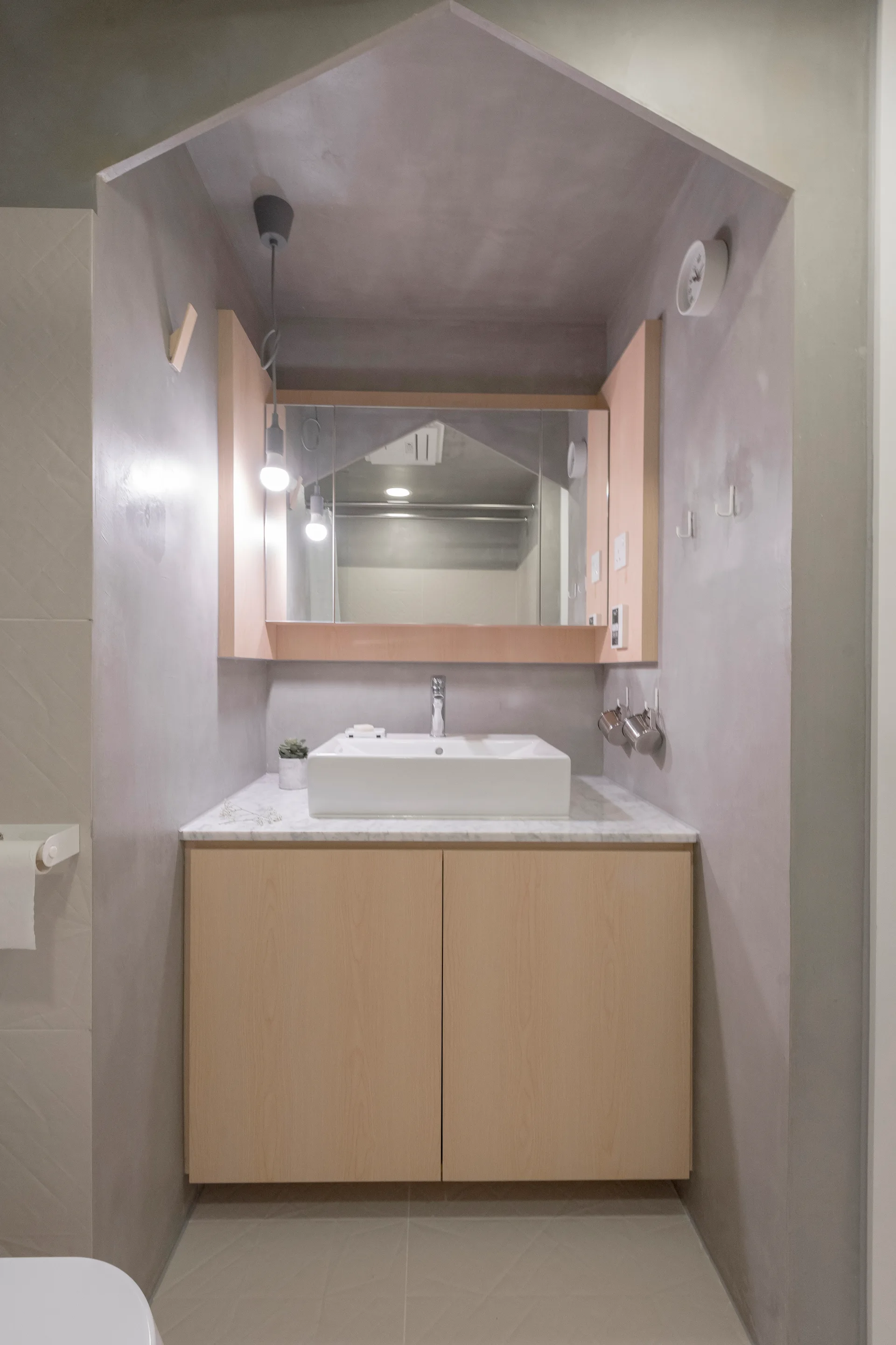 A modern bathroom vanity with a light wood cabinet and white vessel sink set inside a grey plaster alcove with a gabled ceiling structure.