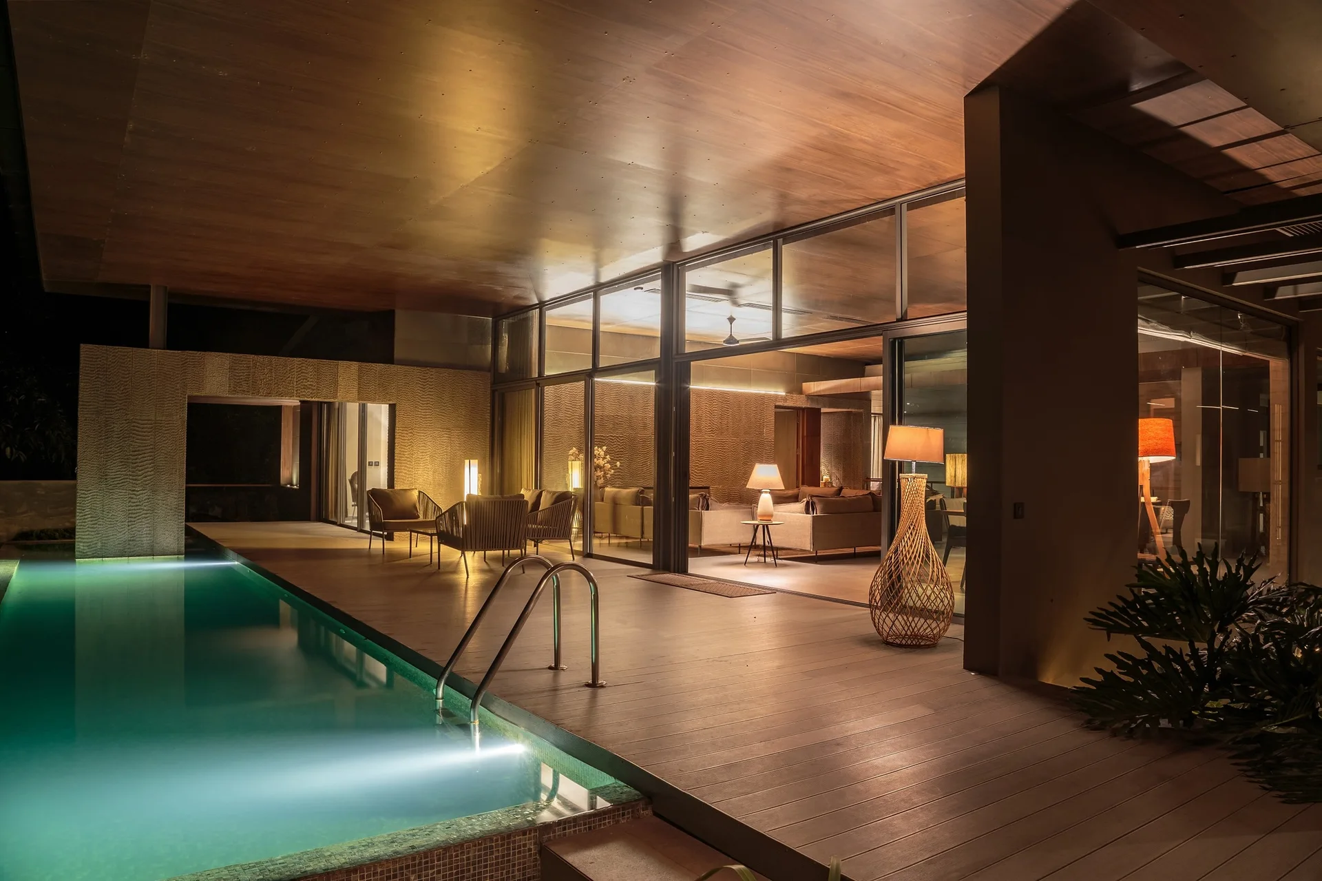 An illuminated swimming pool borders a wooden deck with seating, next to a living room with floor-to-ceiling glass walls and warm lighting.