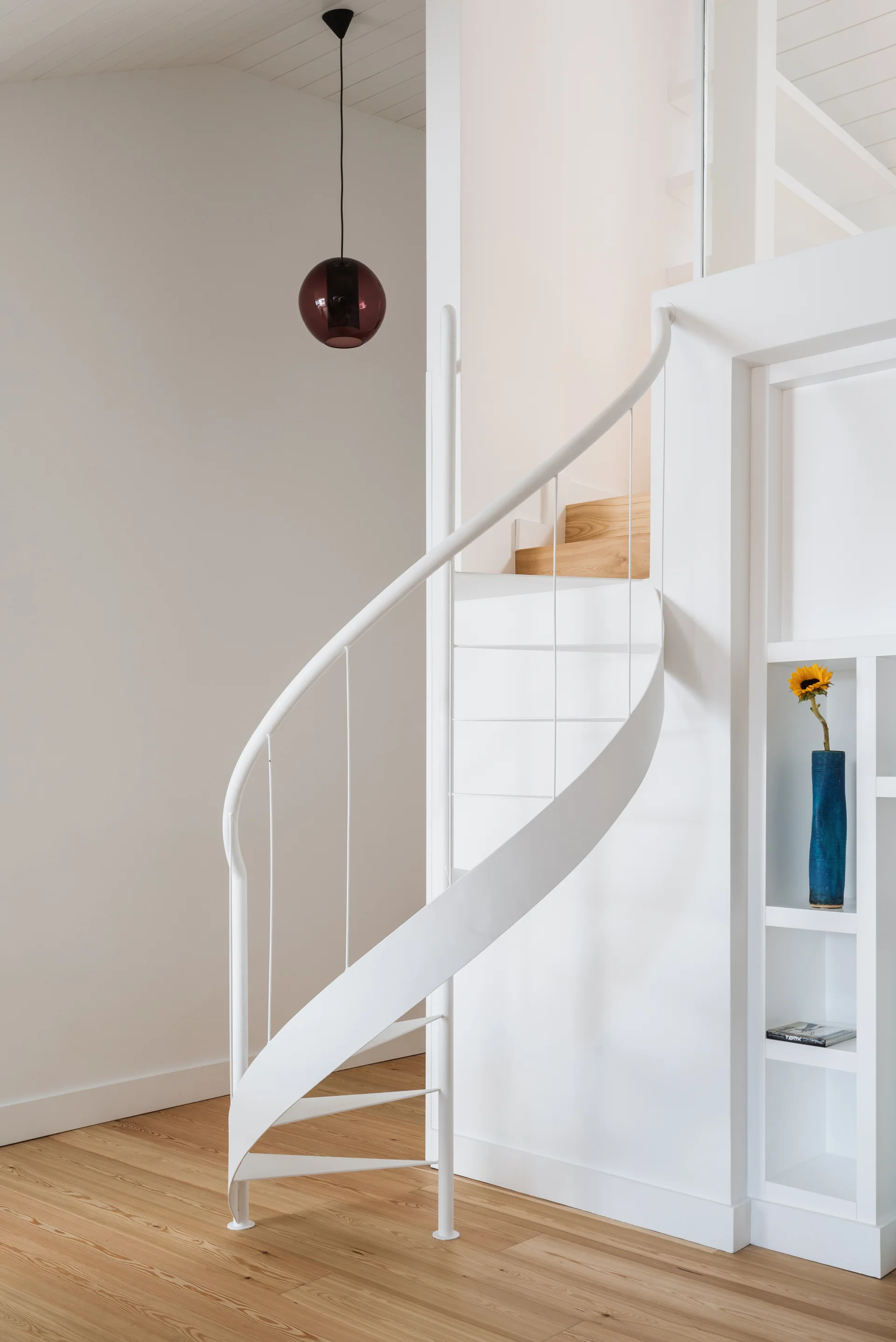 A white metal spiral staircase with light wood treads is shown next to white walls and built-in shelves in a minimalist home.