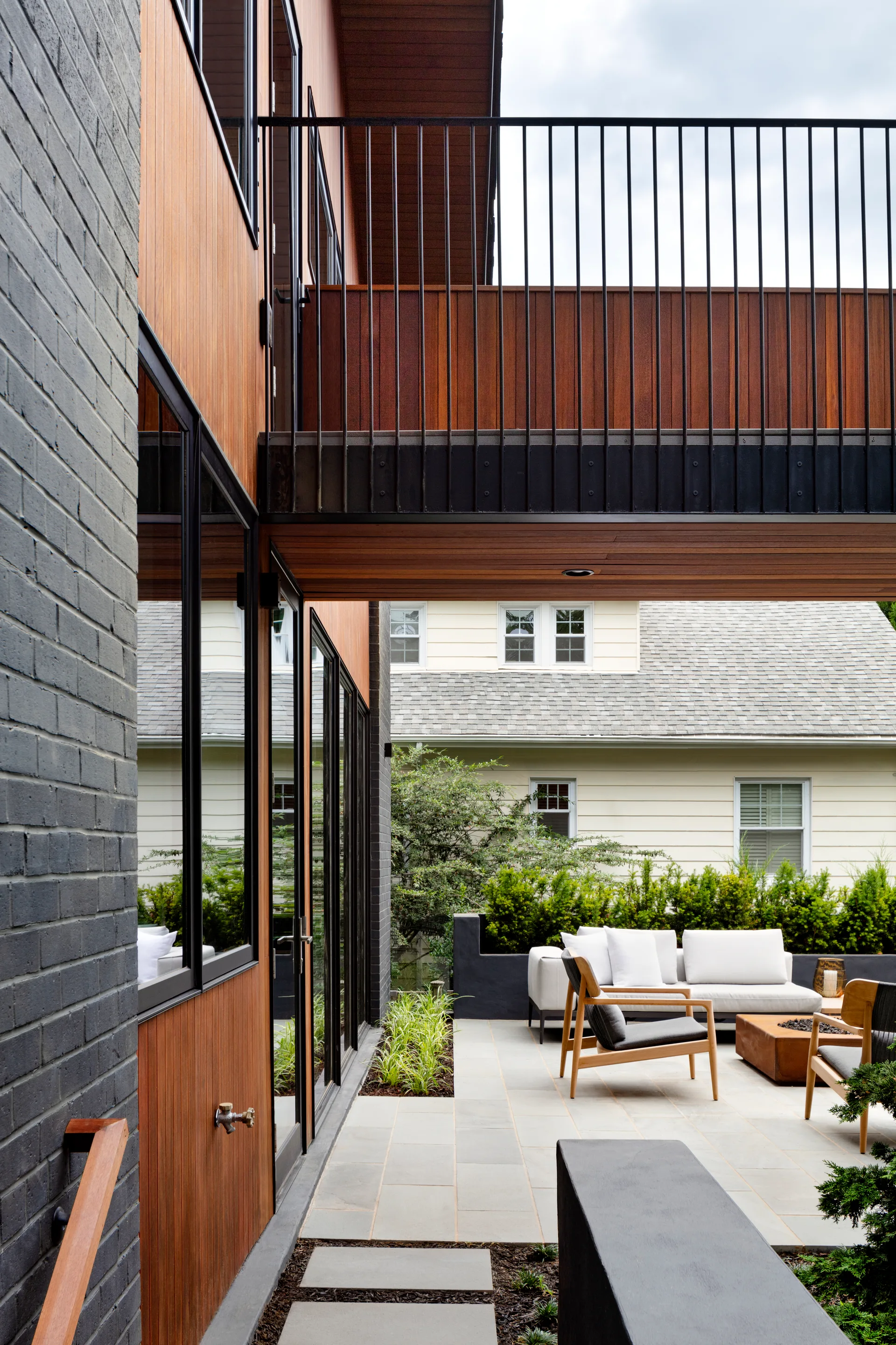 A modern residential patio features stone pavers, a low fire pit, wood-framed chairs, and an overhanging balcony with a black railing.