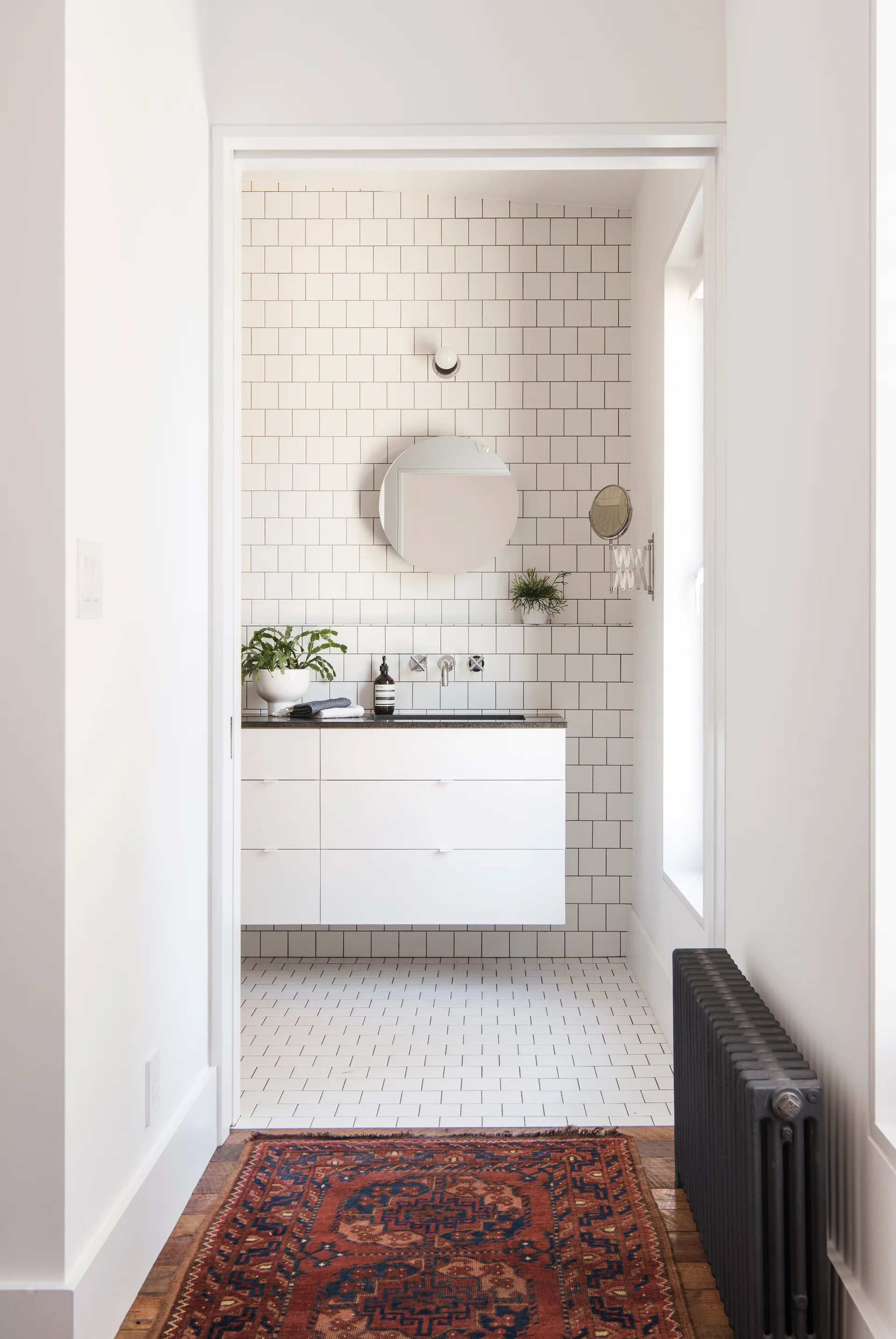 A view through a doorway into a bathroom with white grid wall tiles, a floating white vanity, and a round frameless mirror.