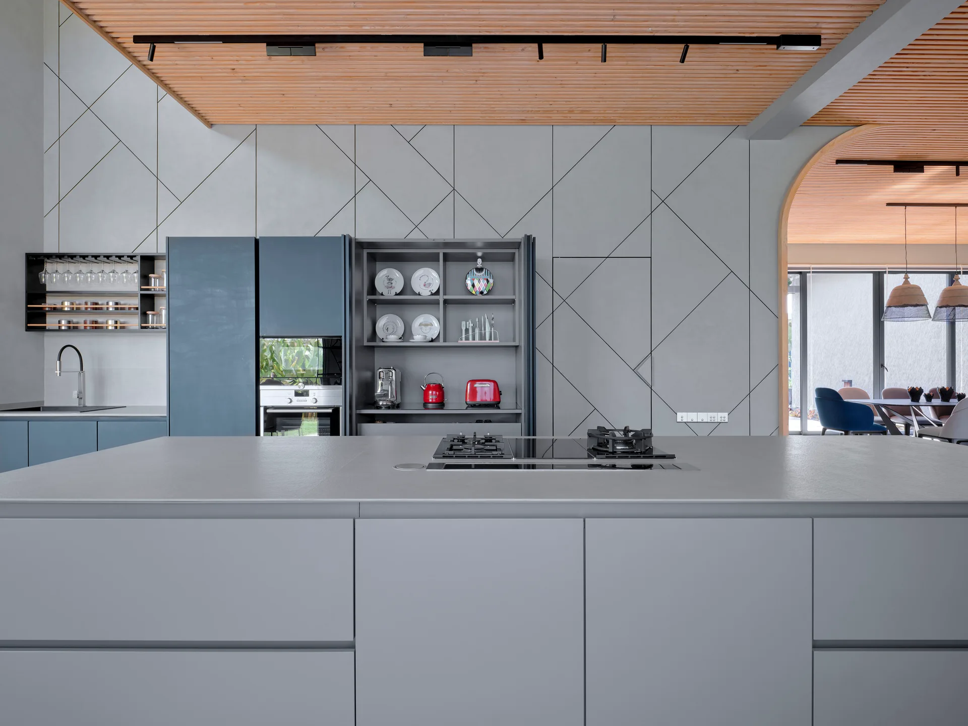 A modern kitchen featuring a gray island with a built-in cooktop, geometric wall panels, blue cabinetry, and a slatted wood ceiling.