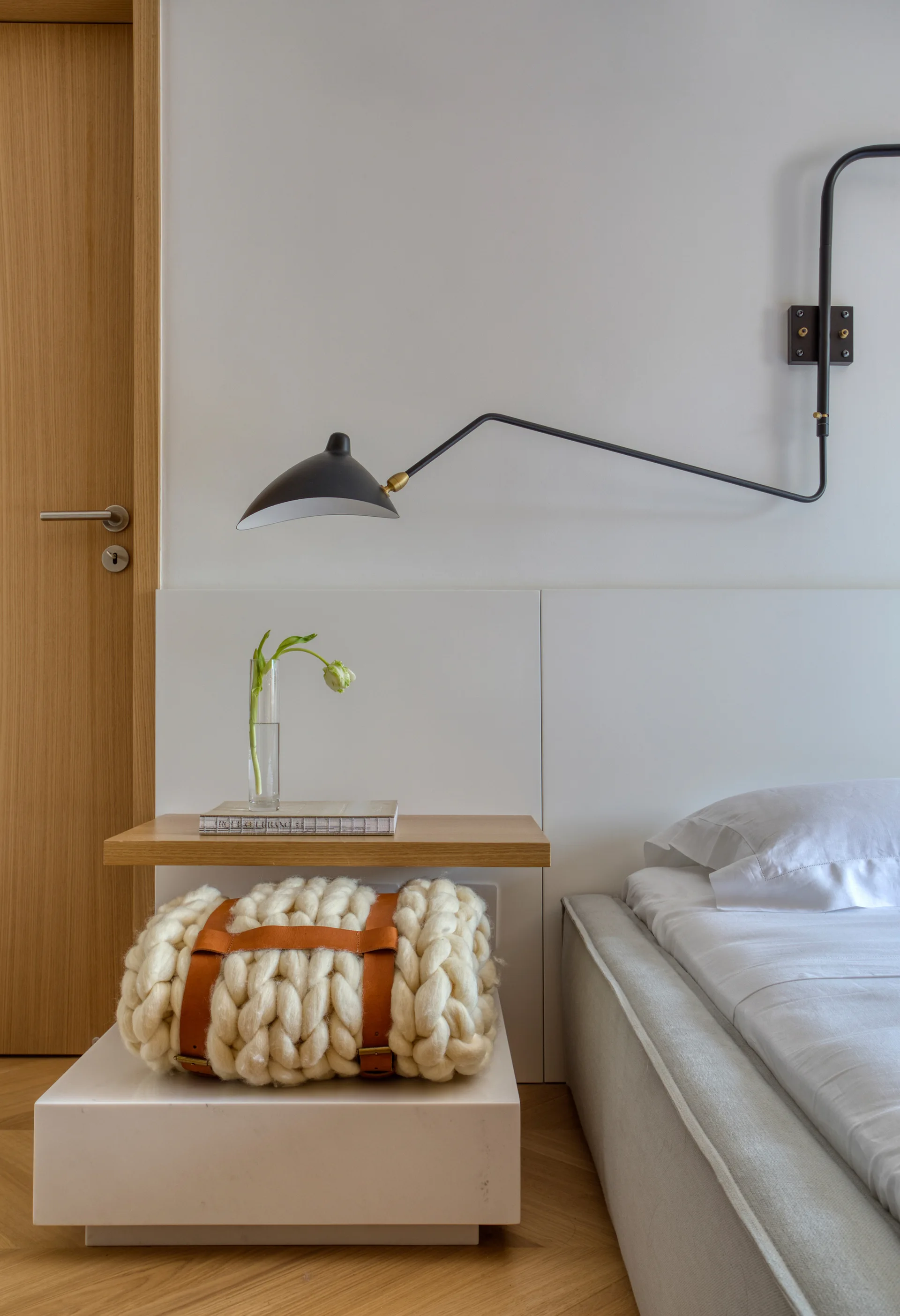 A minimalist bedroom bedside setup featuring a two-tiered nightstand with a wooden shelf, a chunky knit blanket, and a black wall-mounted sconce.