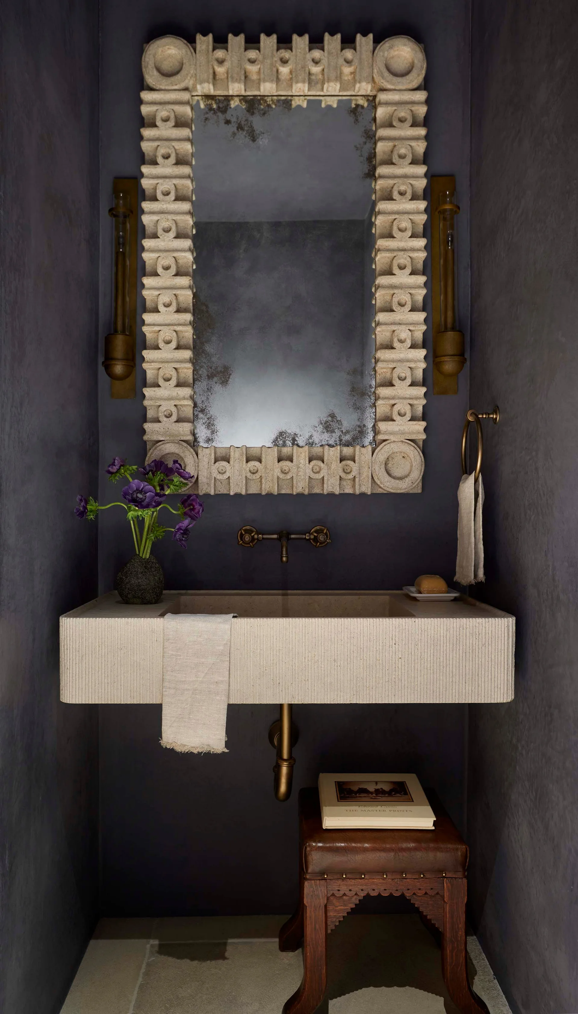 A moody powder room features a wall-mounted fluted stone sink and an ornate stone mirror against textured dark gray walls.