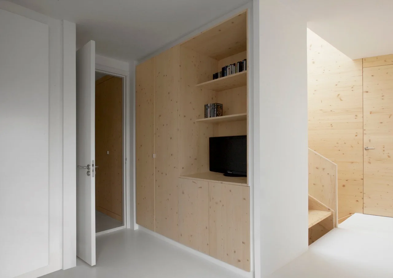 A minimalist white hallway featuring a large light wood built-in storage cabinet with open shelves next to a wooden staircase.