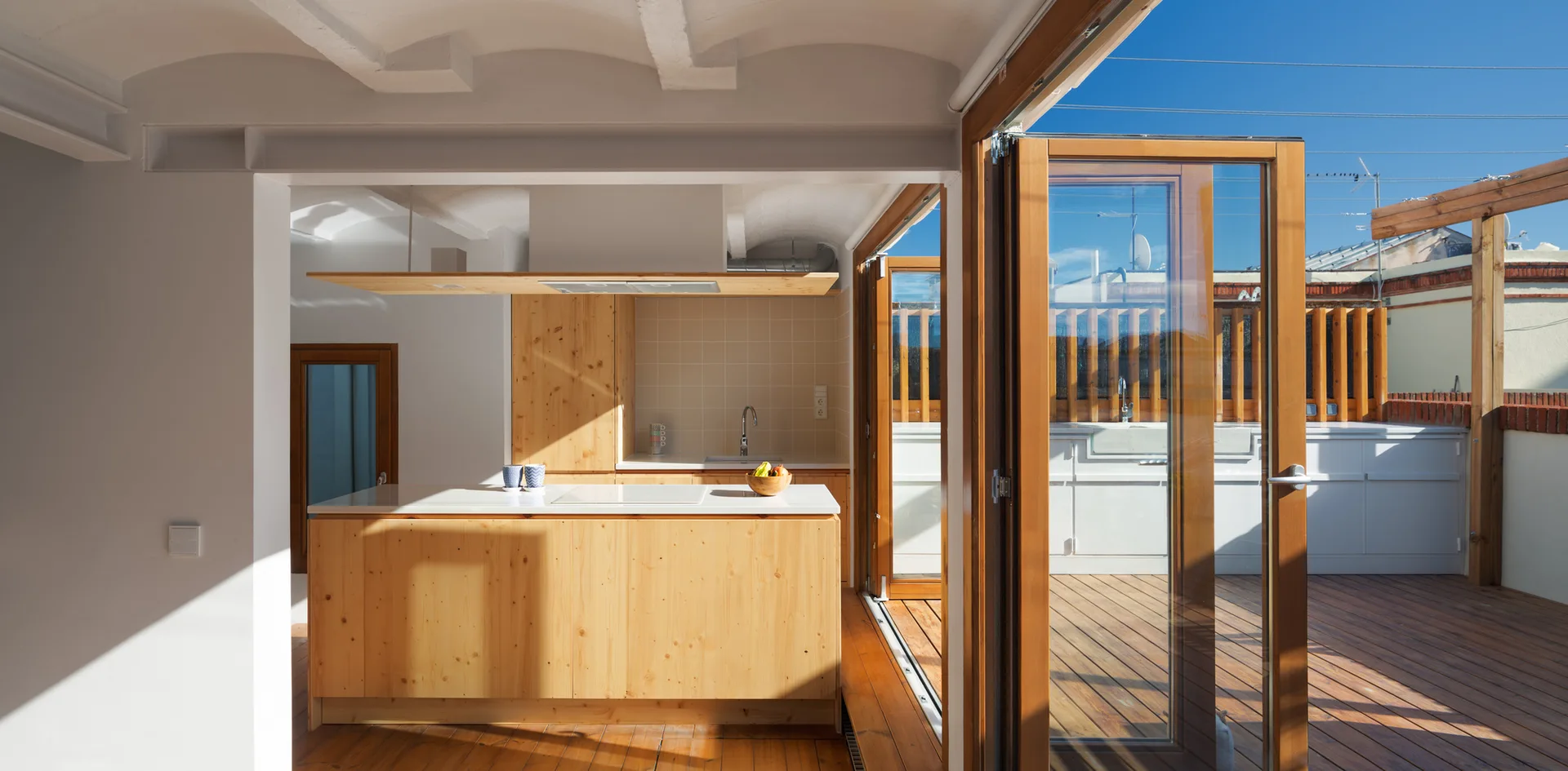 A modern kitchen with light wood cabinets and an island opening onto a sunny wooden terrace through folding glass doors.