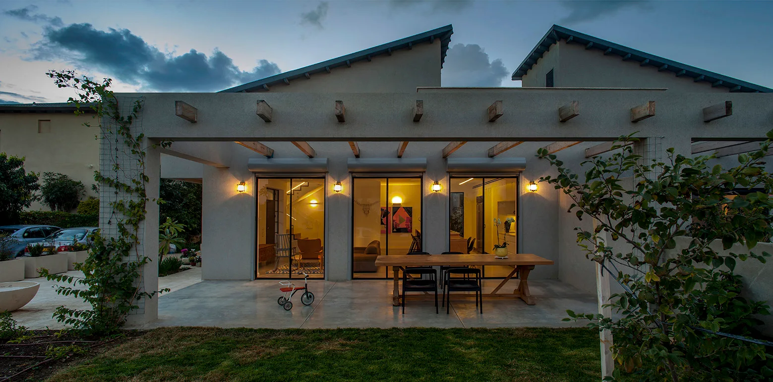 A contemporary residential patio at dusk features a concrete floor, a wooden pergola, and glass sliding doors revealing a brightly lit interior.