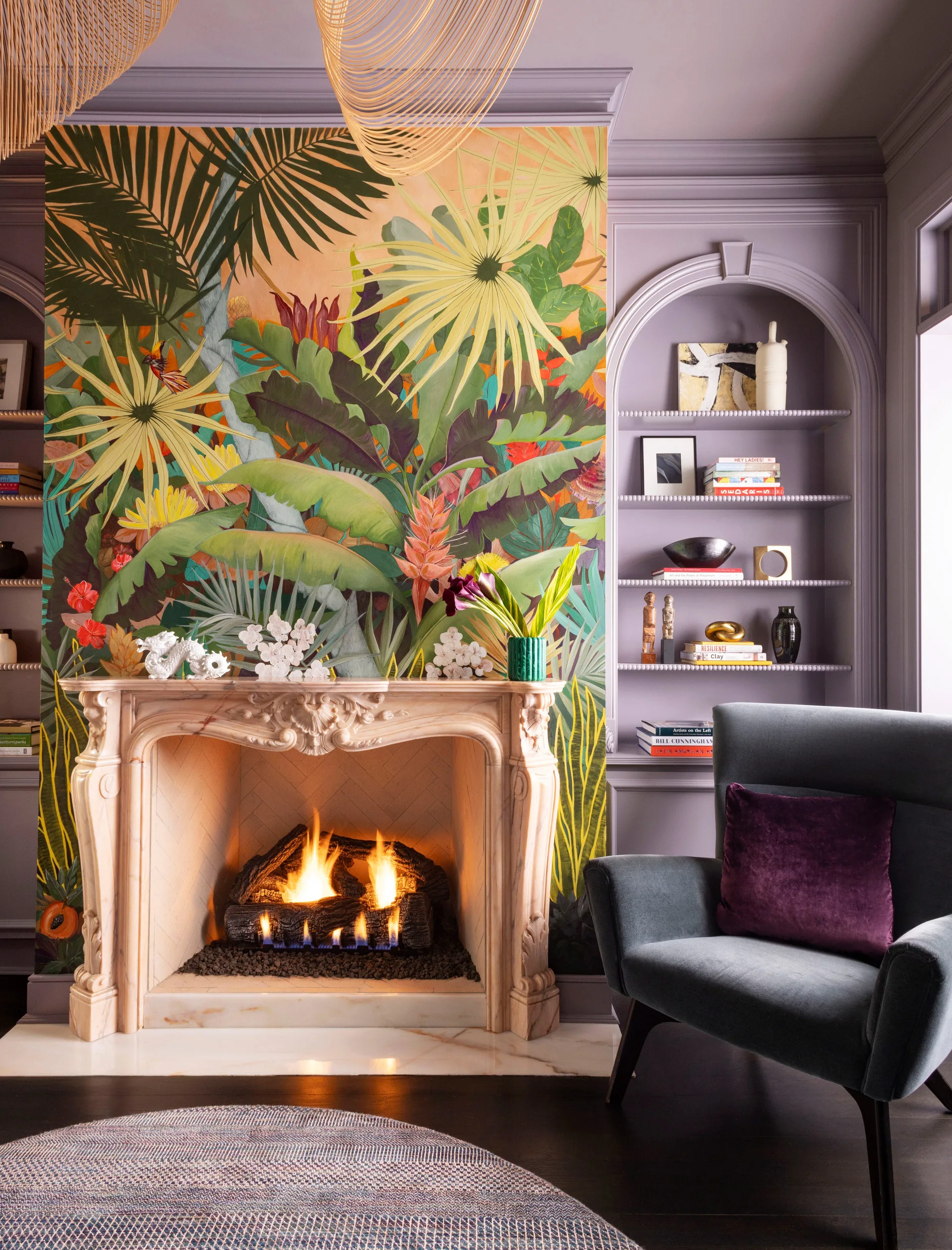 A glowing stone fireplace is framed by a tropical leaf mural and lavender arched built-in shelves next to a velvet armchair.