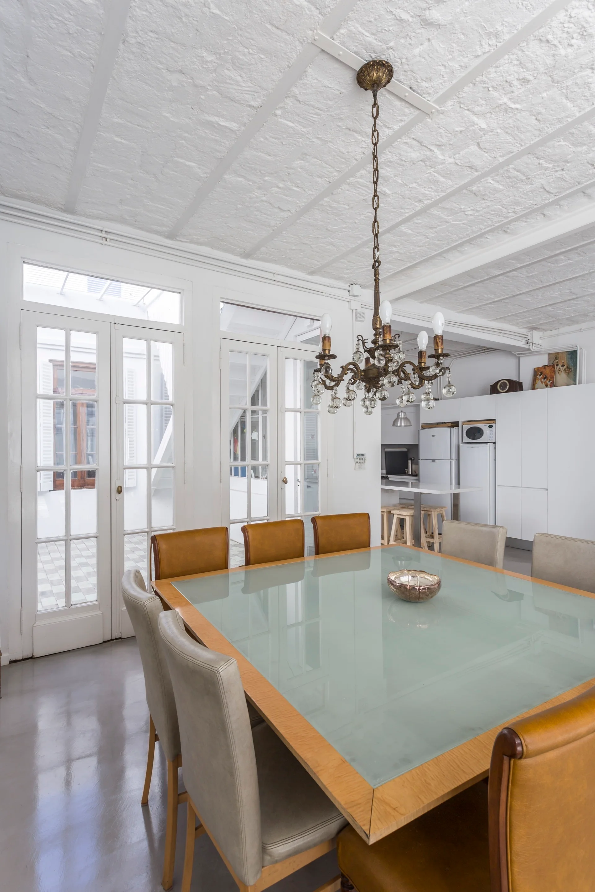 A square dining table with a glass top is surrounded by mixed tan and grey leather chairs beneath an ornate bronze chandelier.