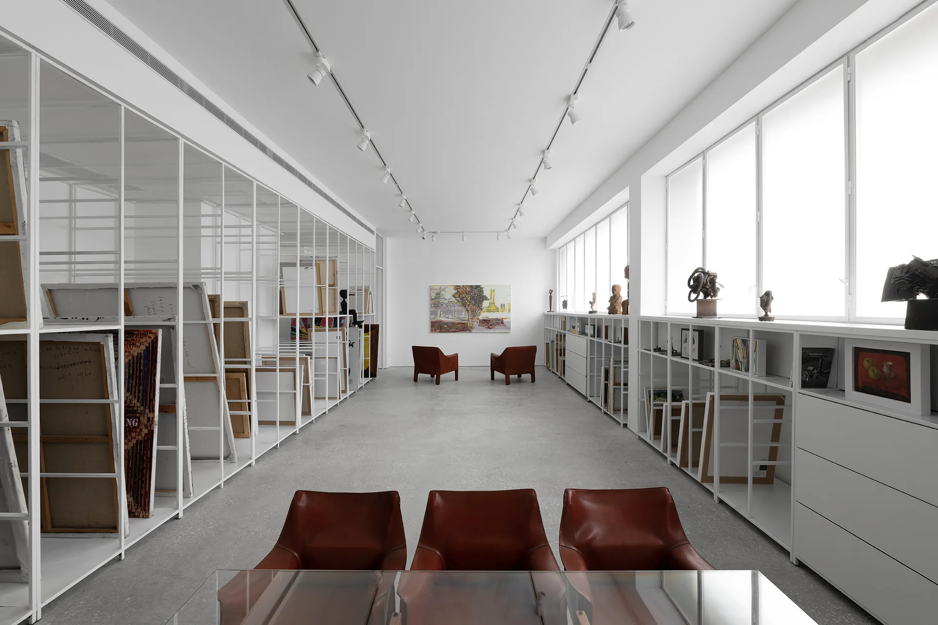 A spacious minimalist gallery features white metal grid shelving, track lighting, concrete floors, and brown leather chairs.