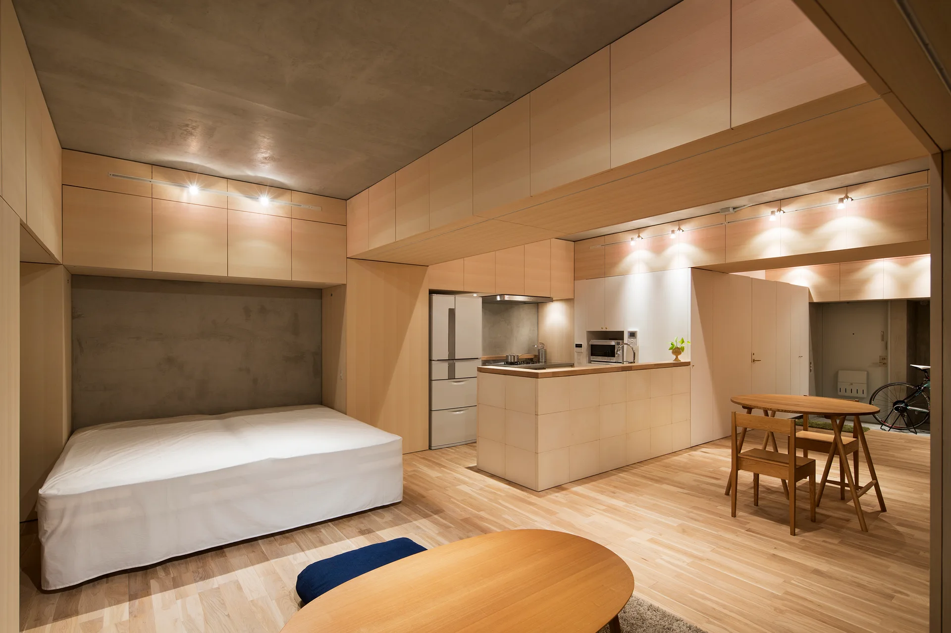 A minimalist studio apartment features an integrated low-profile bed, light wood cabinetry, a kitchen island, and an exposed concrete ceiling.