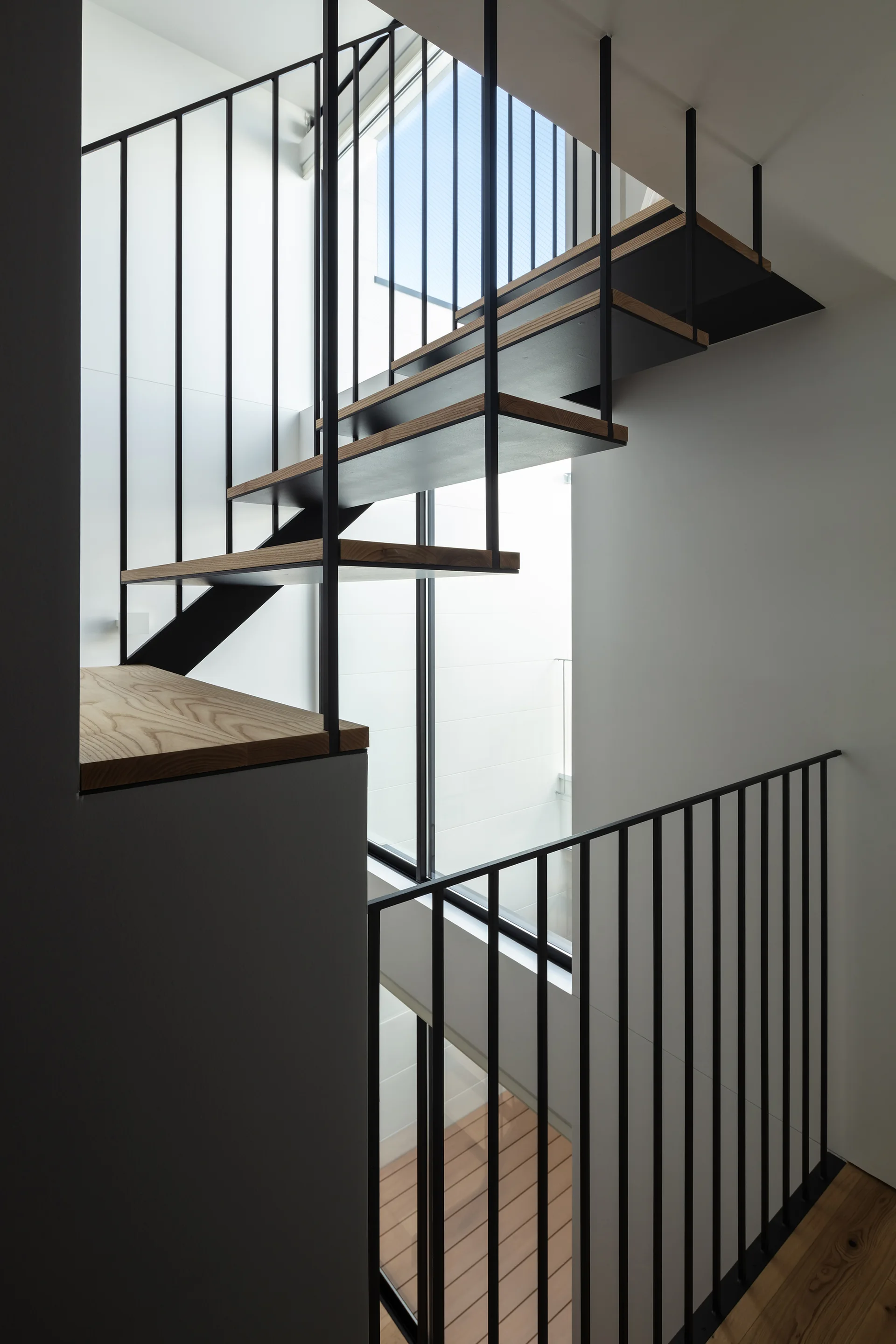 An open riser staircase with light wood treads and a black metal vertical spindle railing rises against white walls next to a tall window.
