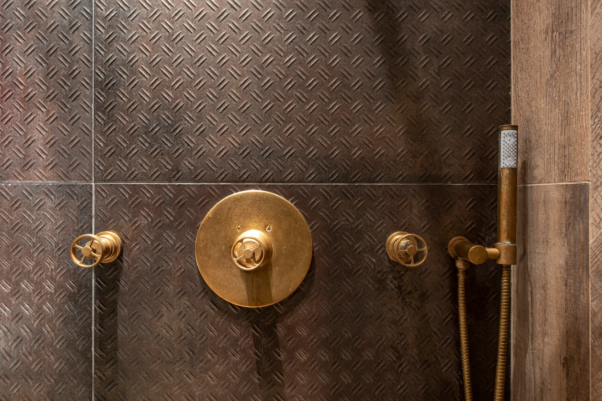 A close-up of dark metallic diamond plate wall tiles in a shower with round brass plumbing fixtures and a handheld wand.