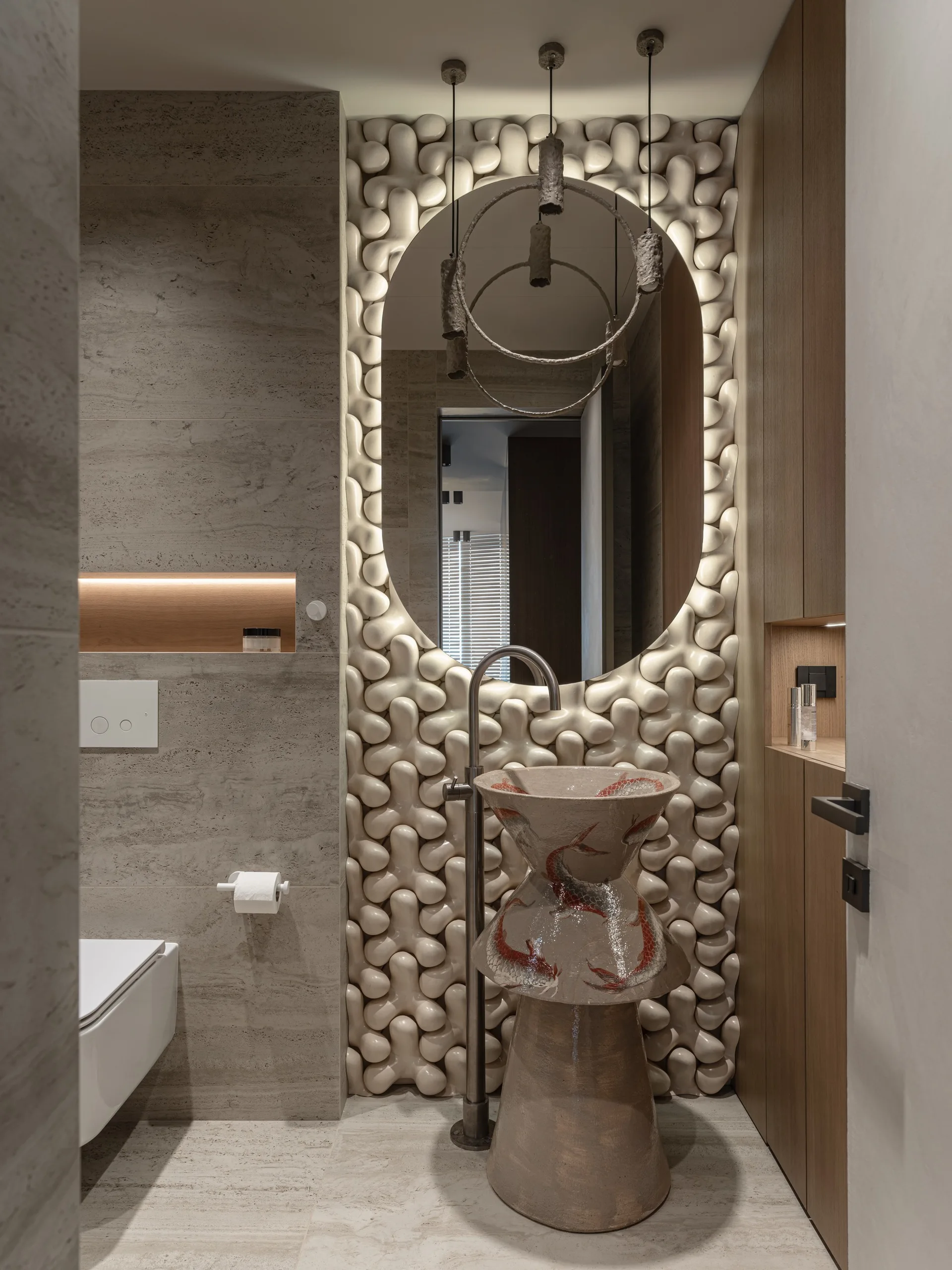 A modern powder room features a sculptural ceramic pedestal sink with fish patterns set against an interlocking 3D tile wall and an oval backlit mirror.