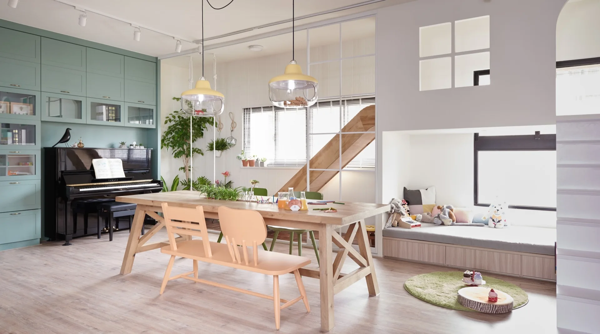 A light-filled family room featuring a light wood dining table, sage green cabinets framing a piano, and a built-in play loft with a window seat.