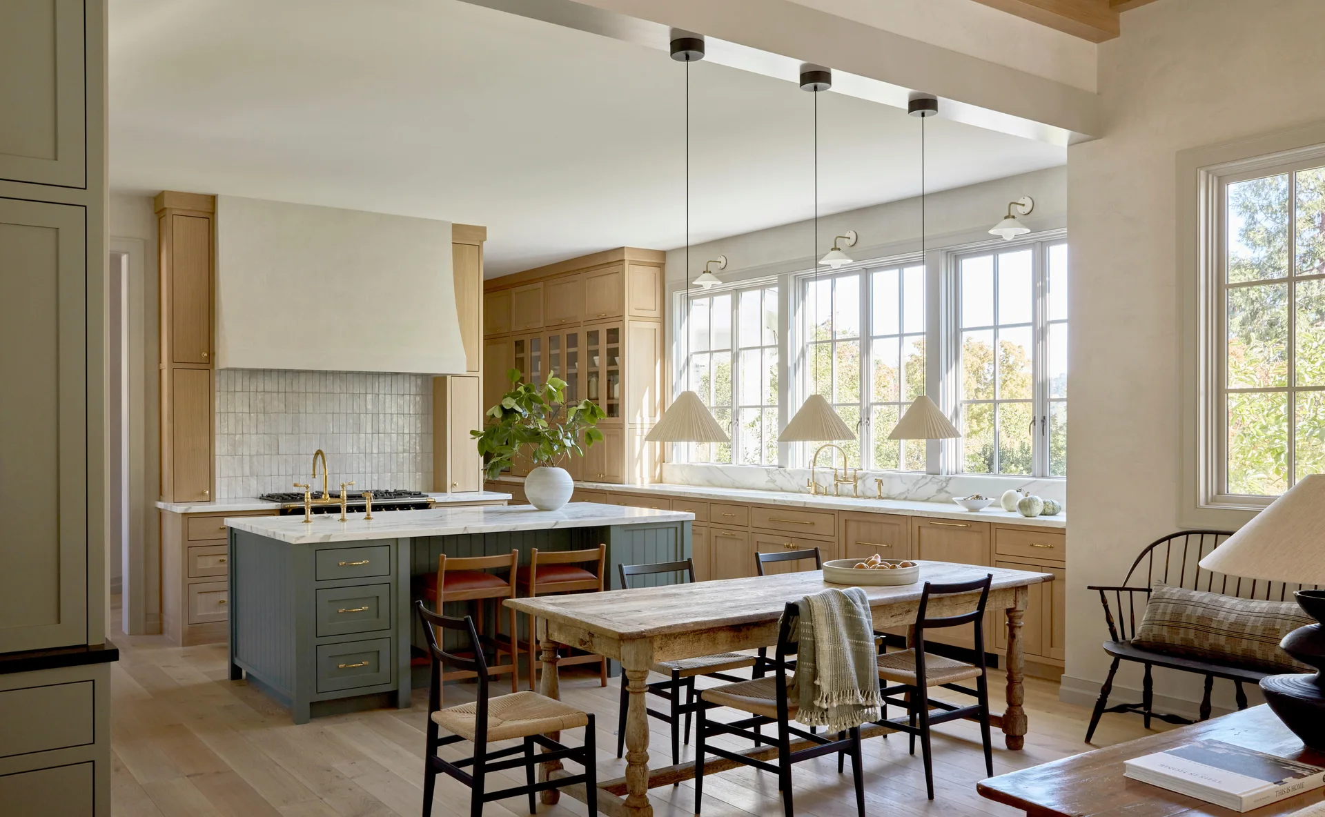 A bright kitchen featuring light wood cabinetry, a sage green island with a marble countertop, and a rustic wood dining table with black spindle chairs.