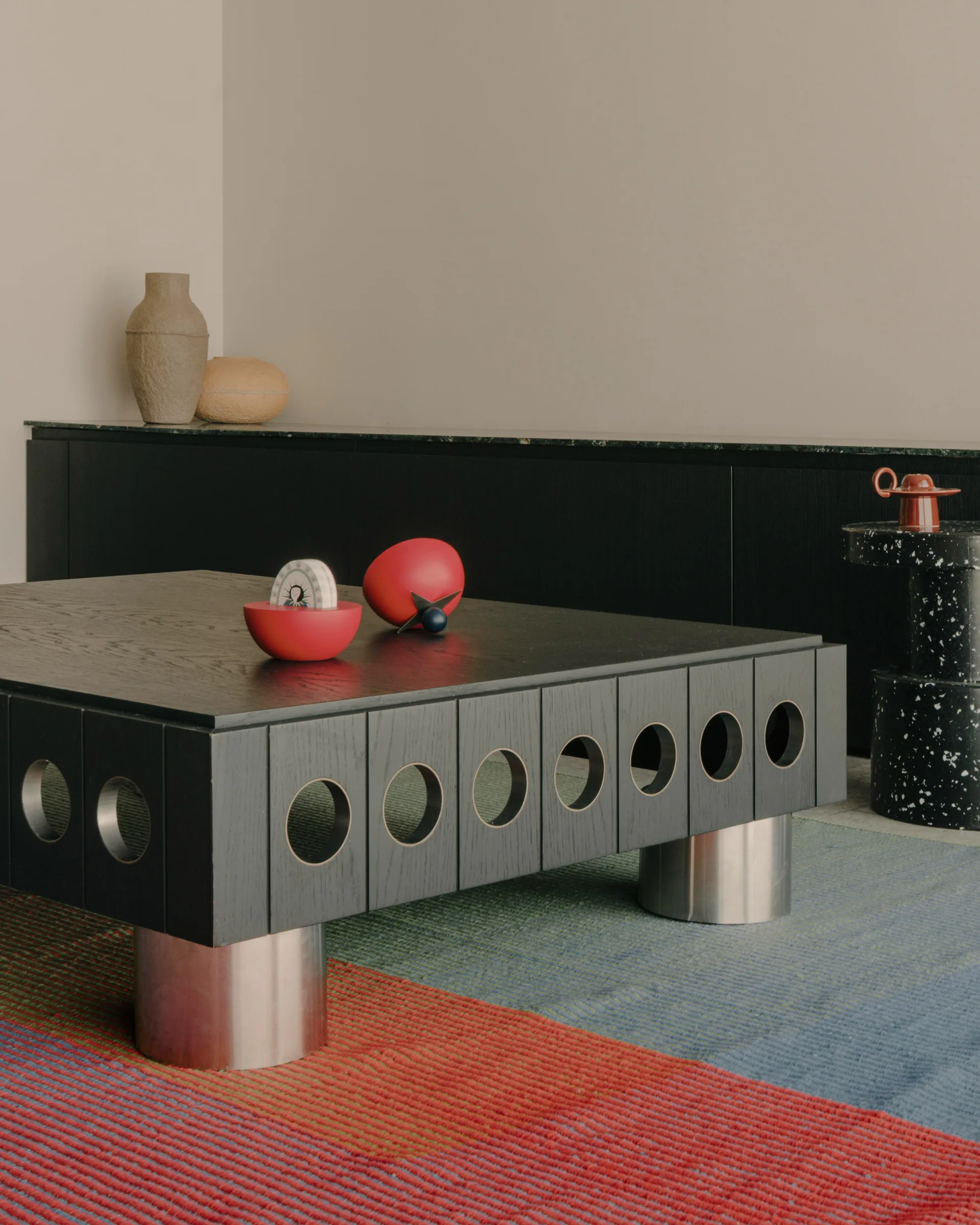 A black wooden coffee table with circular cutout sides and metal cylindrical legs rests on a red and blue striped rug.