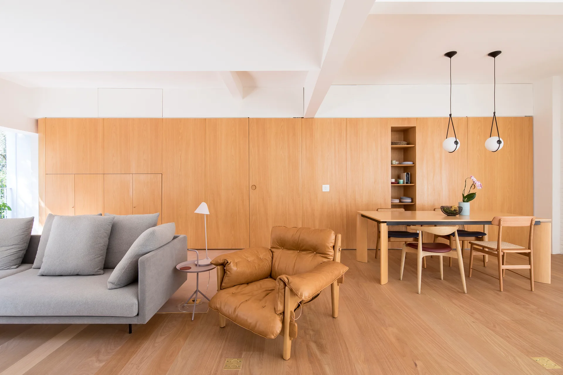 A bright open-plan room features a full-height wood-paneled wall, a grey sofa, a tan leather armchair, and a wooden dining table with chairs.
