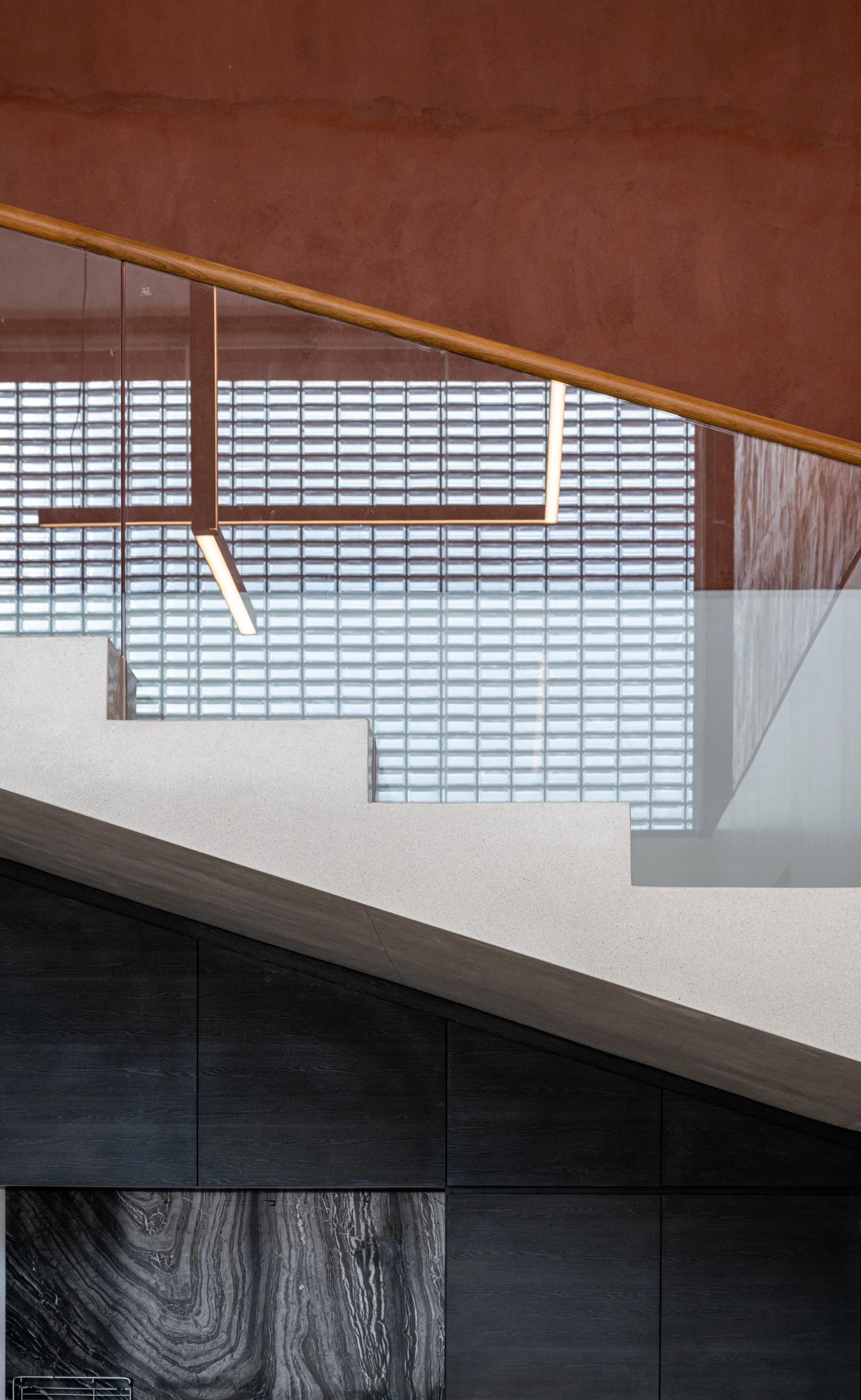 Detail of a concrete staircase featuring a glass railing with a wooden handrail set against a terracotta wall and a glass block background.