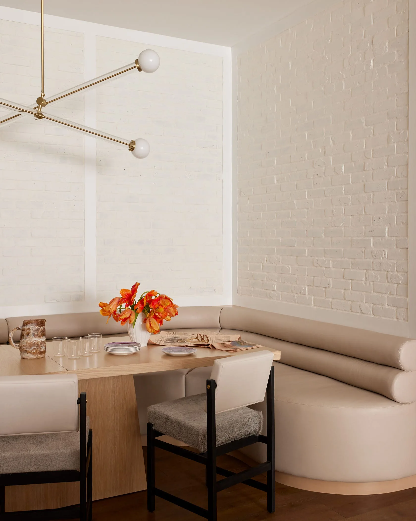 A dining nook featuring a curved beige banquette, a wooden table with chairs, and a modern gold chandelier against painted white brick walls.