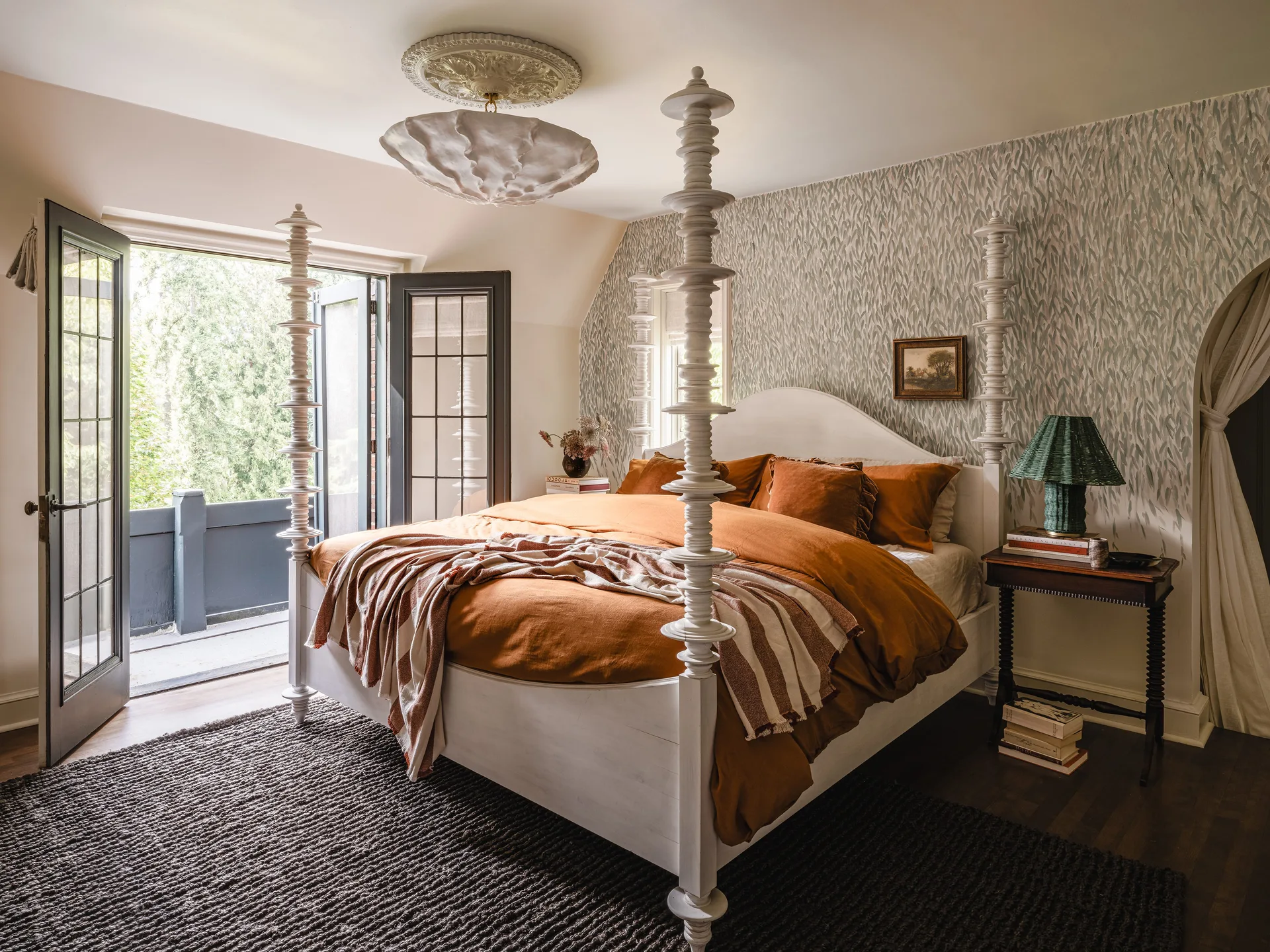A white turned-post bed with rust-colored linens sits against botanical wallpaper next to open French doors leading to a balcony.
