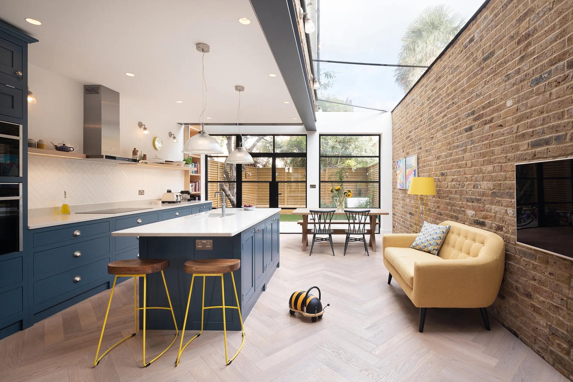 An open-plan kitchen features navy blue cabinetry, a central island, a yellow sofa against an exposed brick wall, and a sloped glass skylight.