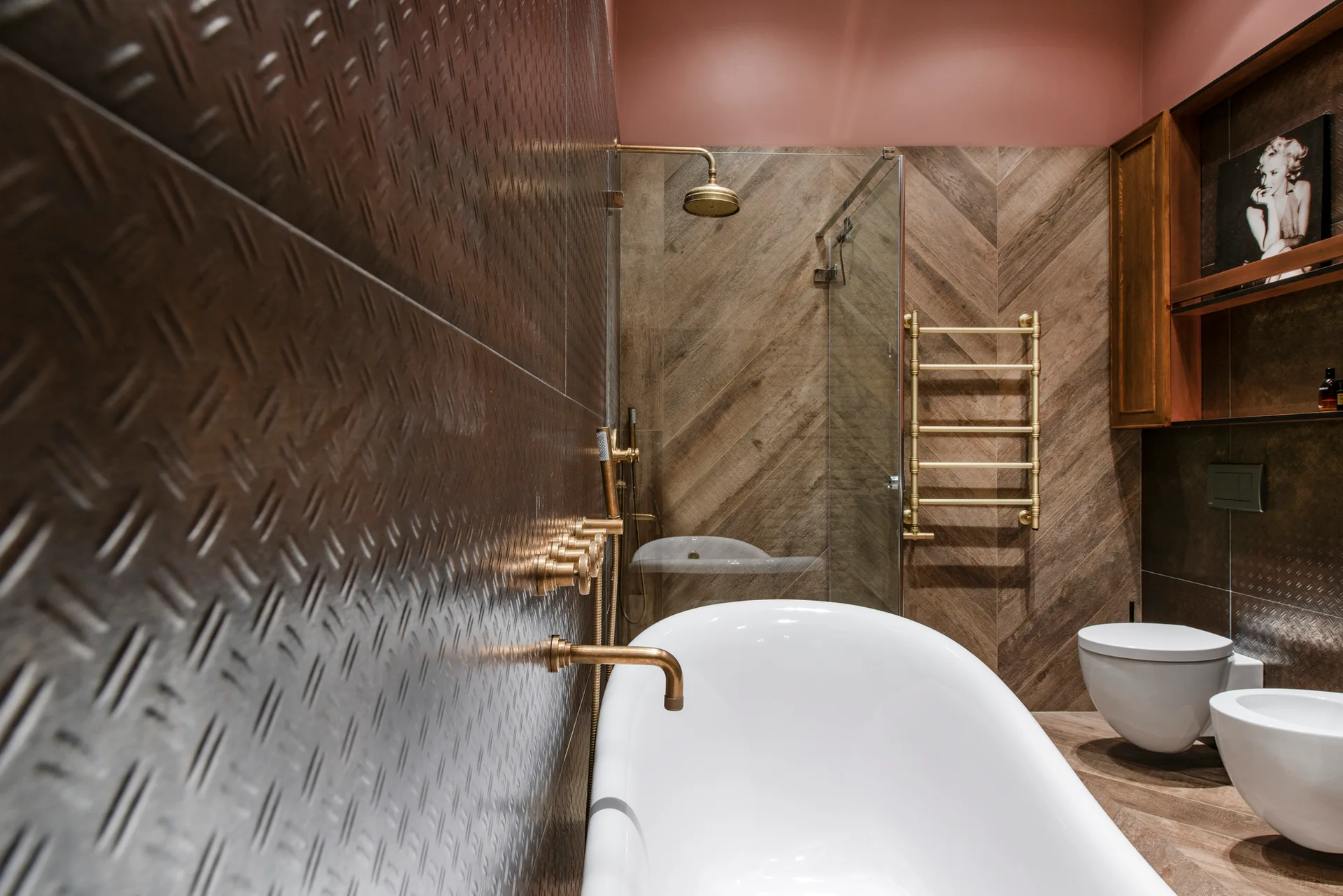 A modern bathroom features a freestanding white bathtub, brass fixtures, a diamond plate wall, and herringbone-patterned wood-look tiles.