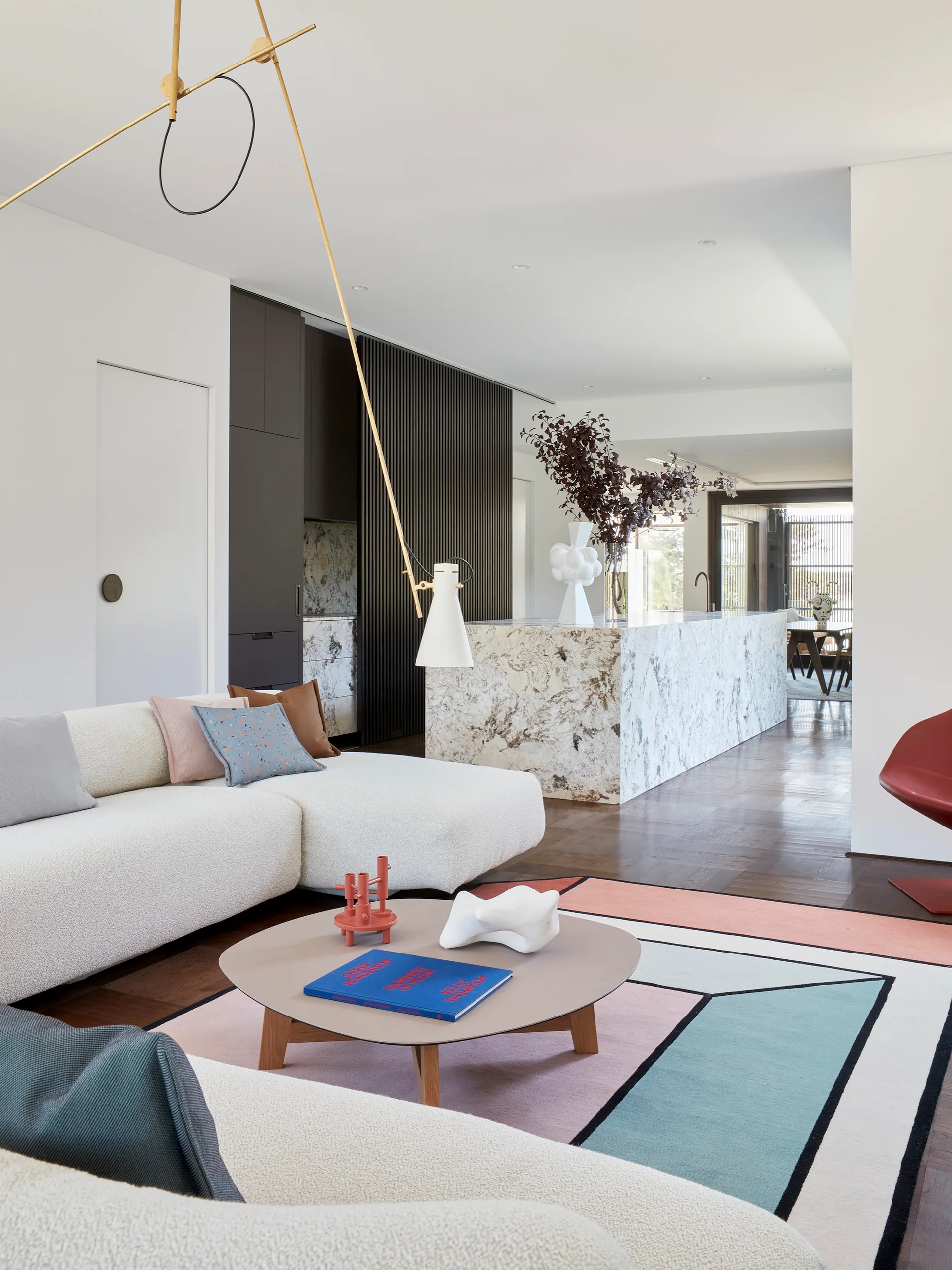 A modern open-plan living room features a cream textured sectional sofa on a geometric block-patterned rug next to a marble kitchen island.