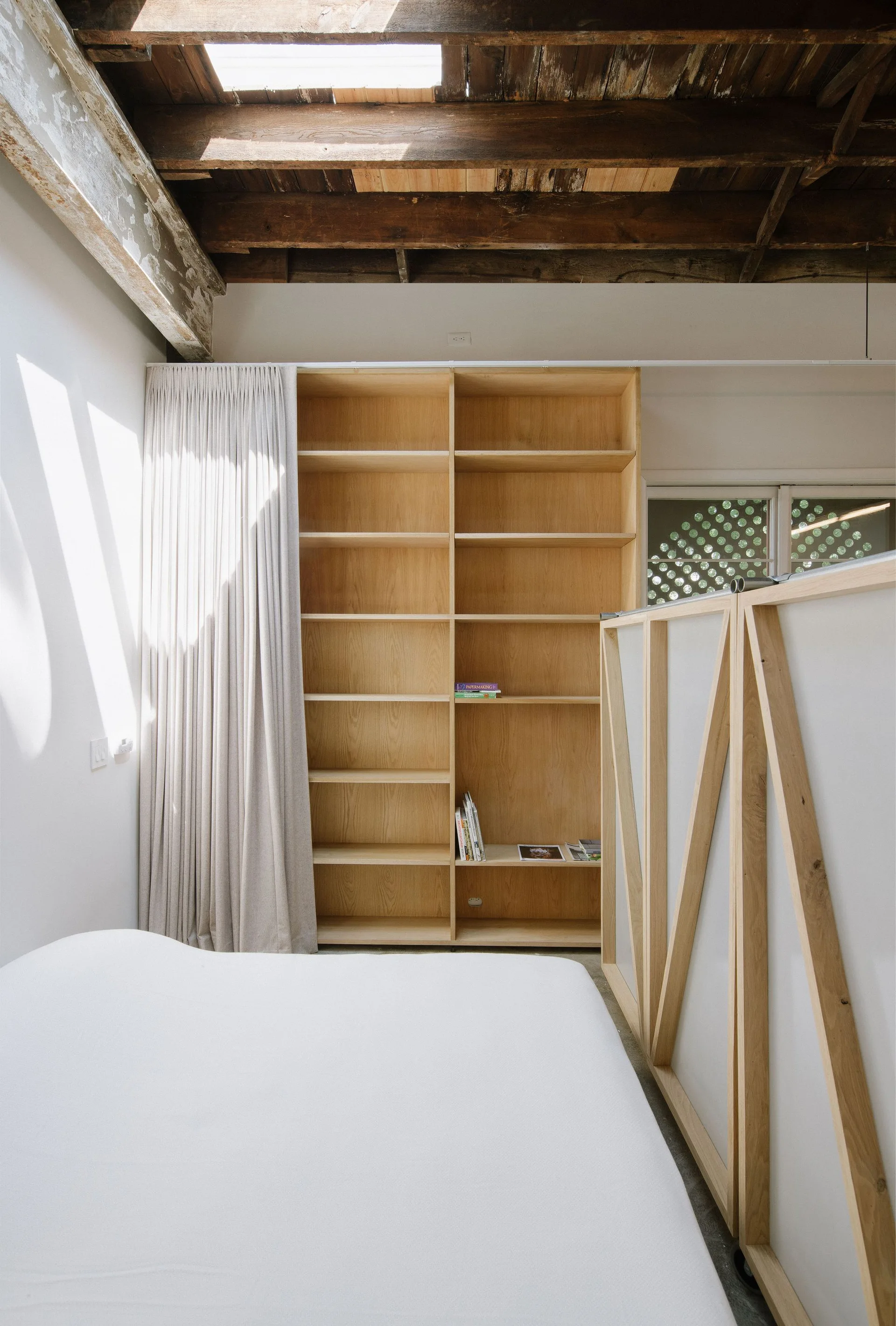 A loft bedroom features a large light wood bookshelf, a custom wood room divider, a white bed, and exposed ceiling beams with a skylight.