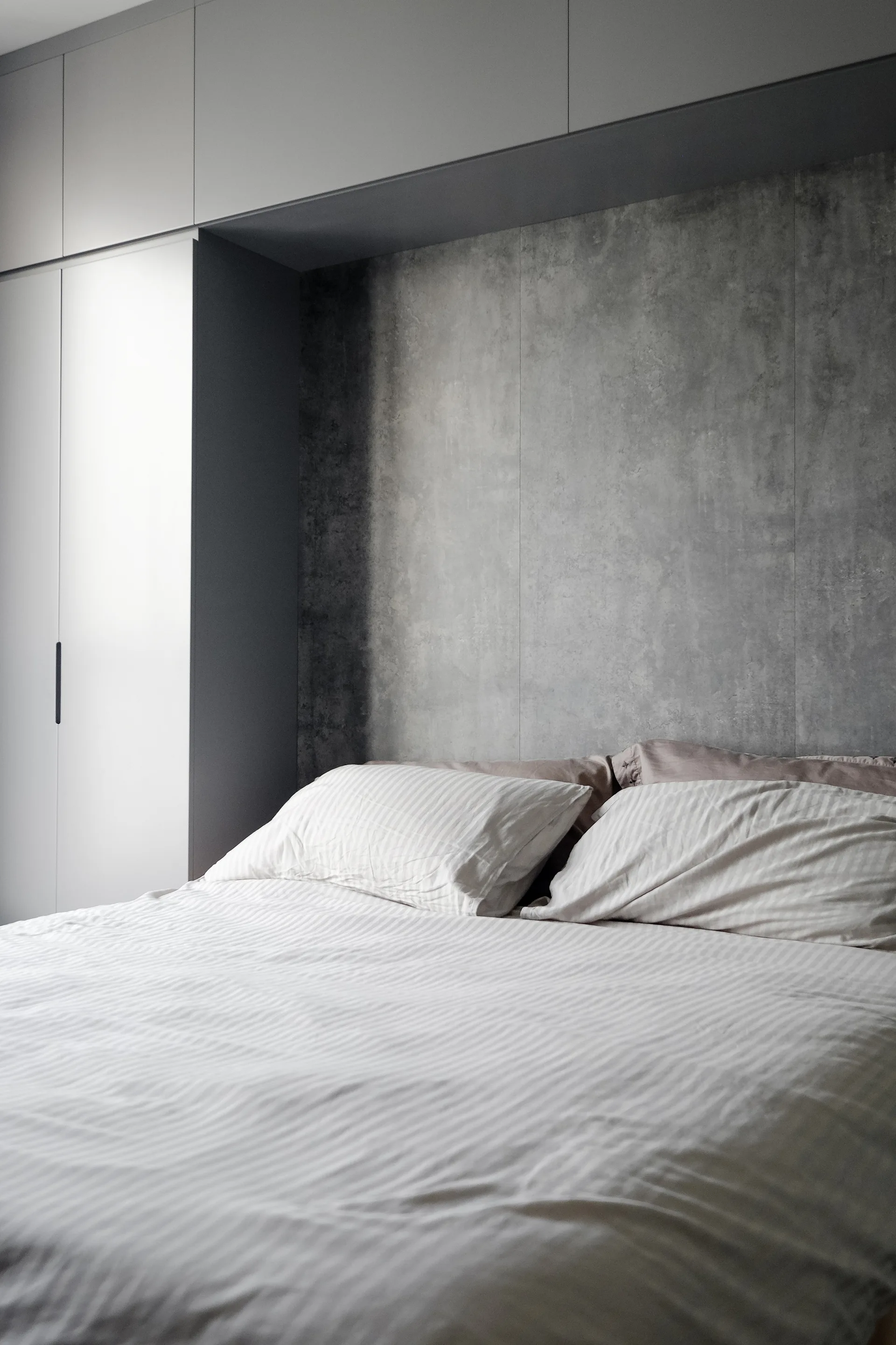 A bed with striped bedding sits against a textured gray concrete wall surrounded by matte gray built-in wardrobes.