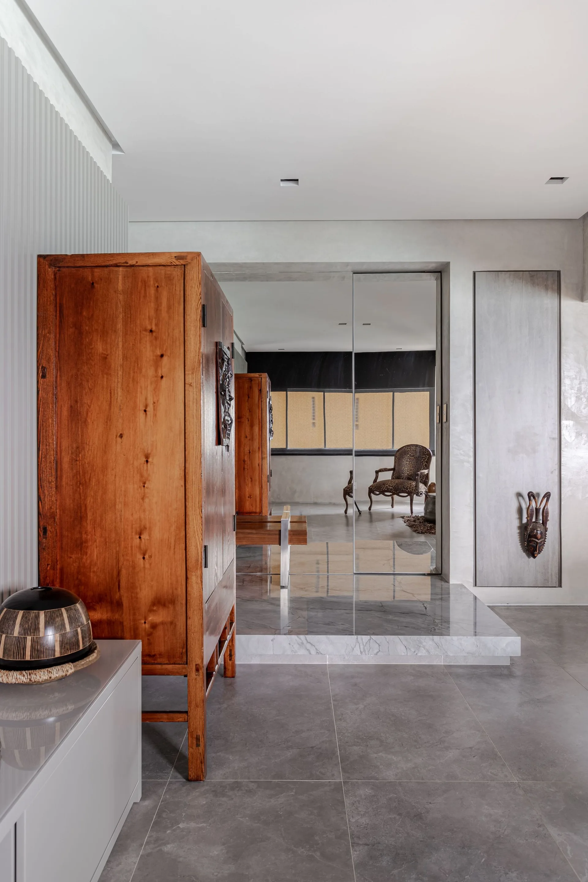 A contemporary hallway features a large wooden armoire next to a raised stone platform and a tall mirrored wall reflecting a patterned armchair.