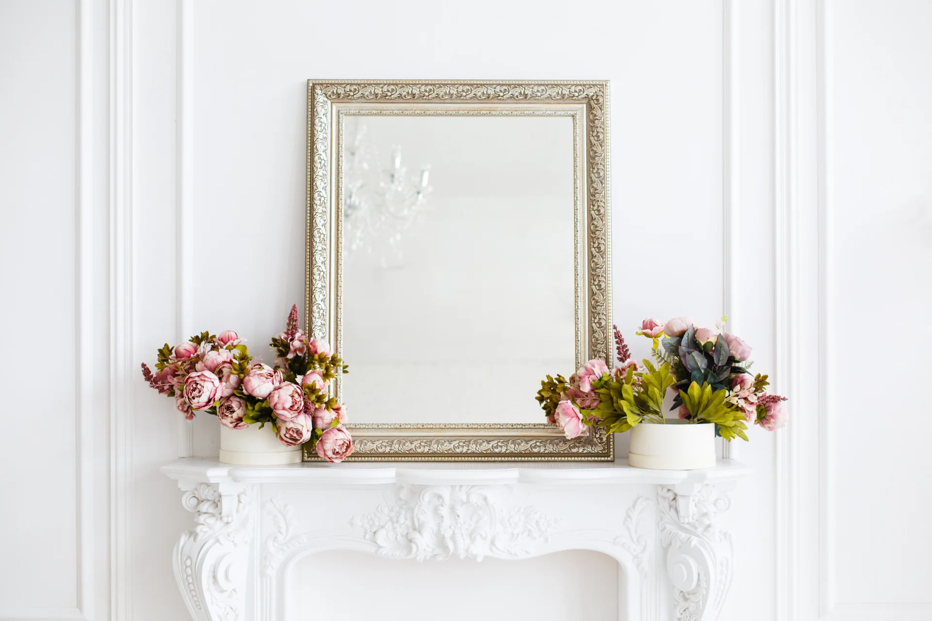 A silver ornate mirror sits on a highly detailed white fireplace mantel, flanked symmetrically by two pink floral arrangements in round cream boxes.